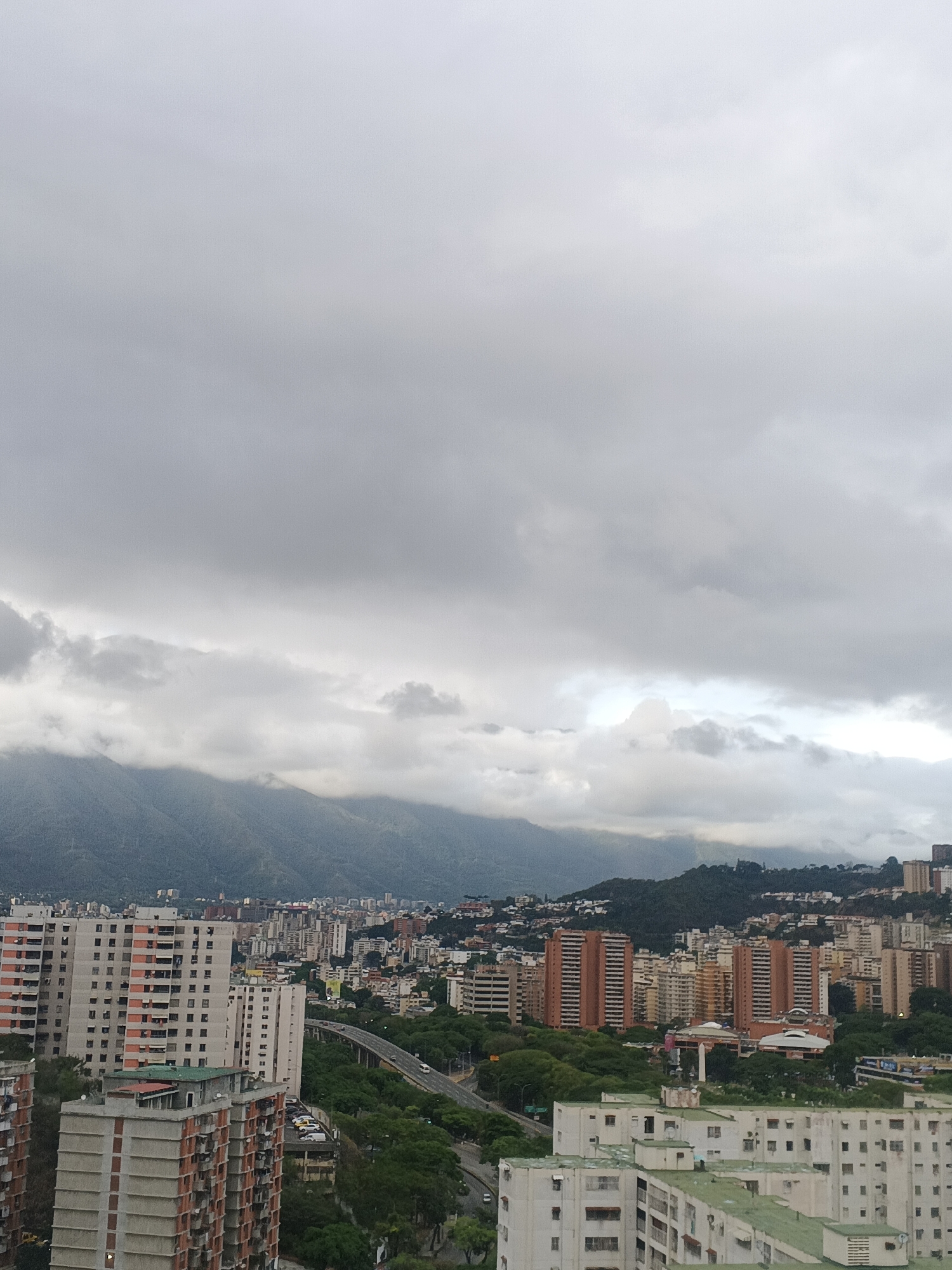 This is an eye-level, outdoor shot of a portion of Caracas, Venezuela on an overcast day. The foreground shows densely populated residential neighborhoods with high-rise apartment buildings of varying heights and colors, primarily cream, orange, and white. A tree-lined highway cuts through the urban landscape.
In the mid-ground, additional buildings stretch toward the base of a mountain range that is partially obscured by low-hanging clouds and mist. The sky above is uniformly gray, filled with dense cloud cover.