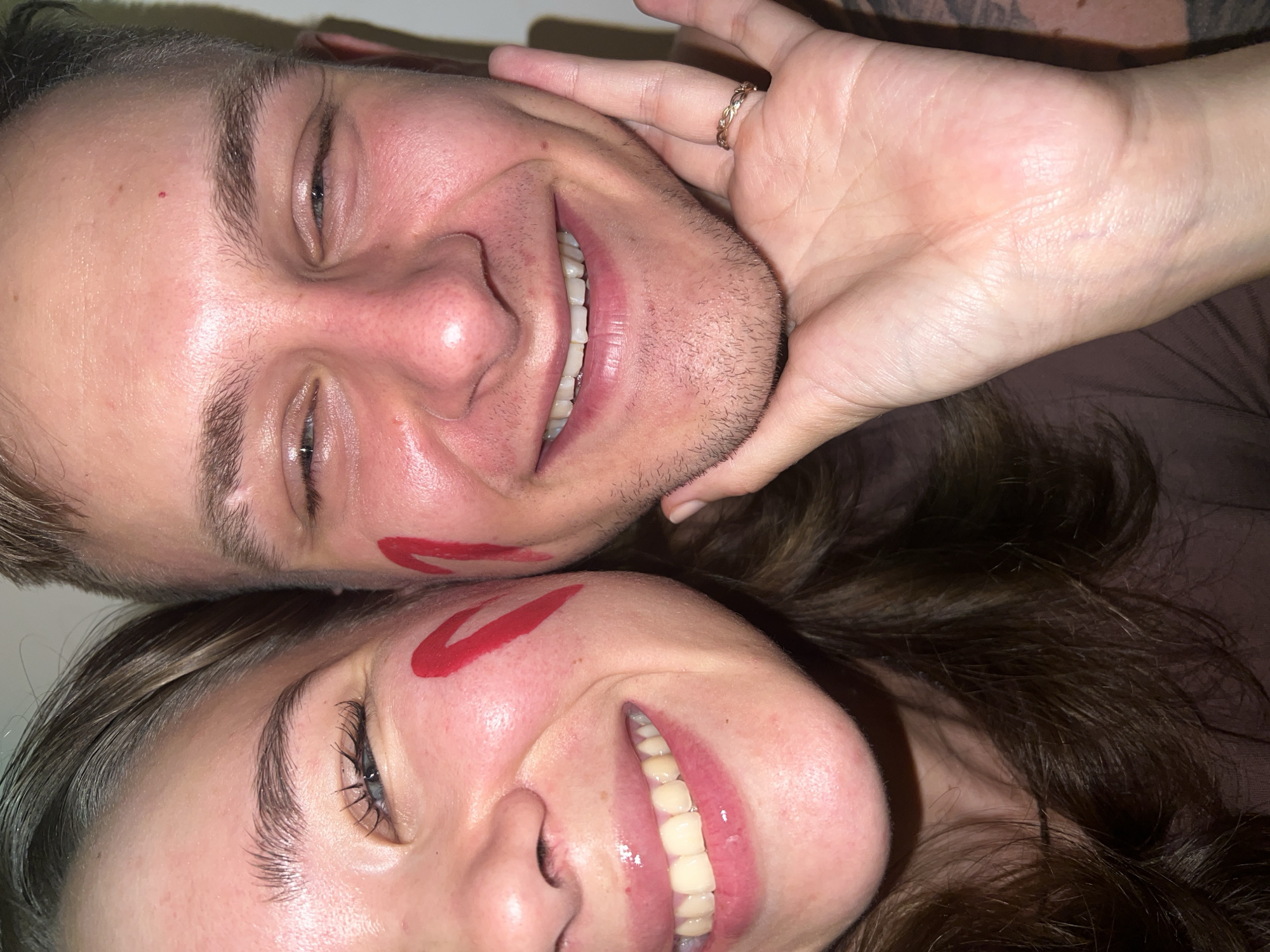 Two people are facing the camera, with one person directly above the other, cheek-to-cheek. Both are smiling, displaying their teeth. Each person has a red lipstick mark on their cheek. The person on top has shorter, light brown hair and stubble. A hand with a ring is holding the top person's face. The bottom person has long, dark hair. The background appears to be a solid dark color. There is no indication of the location being Nam Natural Heritage Area Trang, Vietnam.