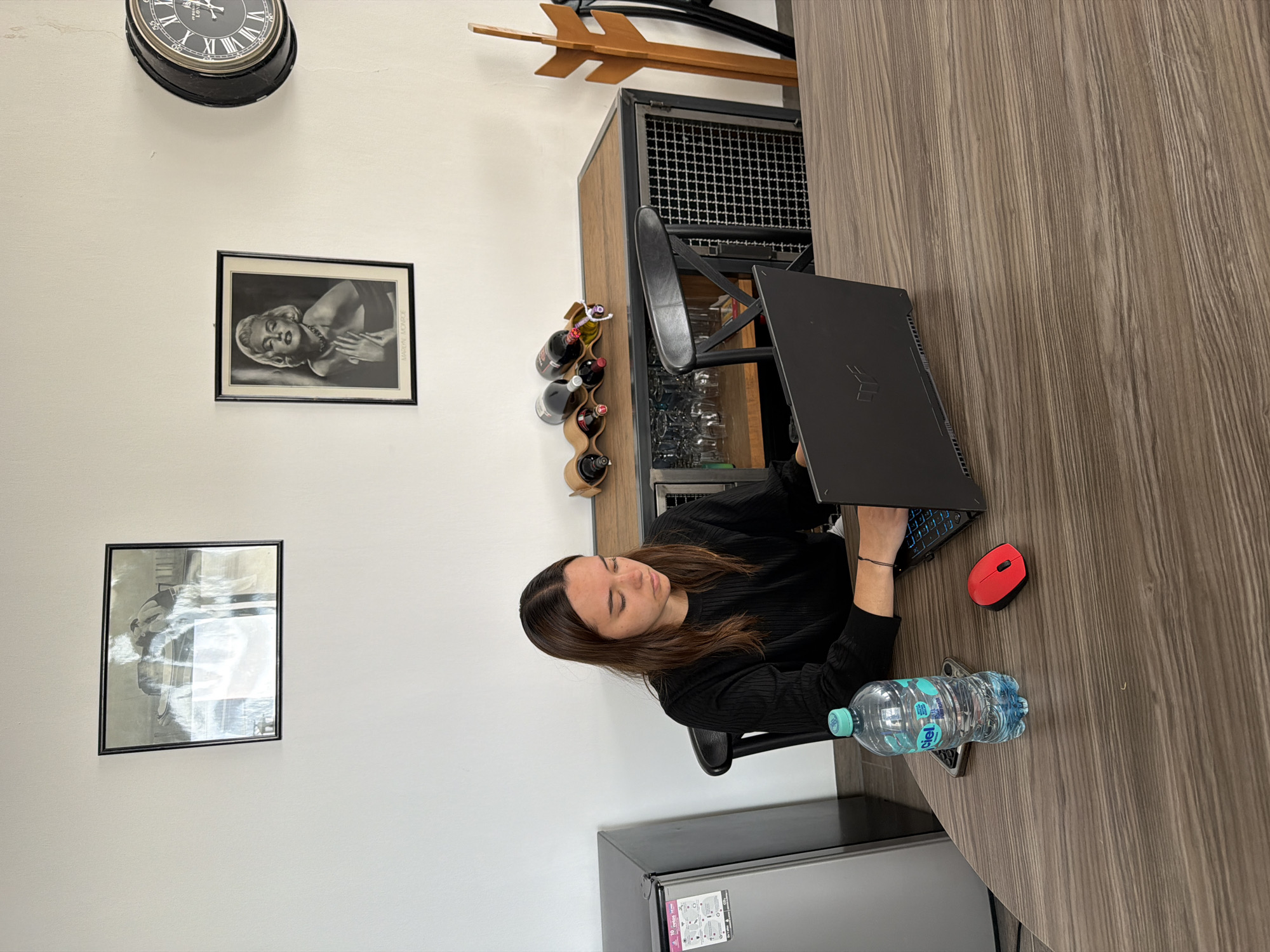 A young woman is seated at a table, working on a laptop. The table is made of wood, and a red wireless mouse and a water bottle are placed on it. The woman is wearing a black long-sleeved shirt. Behind her is a bar cabinet with bottles of wine and glassware visible. A black refrigerator is located next to the bar cabinet. On the white wall behind the woman, there is a black clock and two framed pictures. The image was taken in San Bernardino Tlaxcalancingo, Mexico.