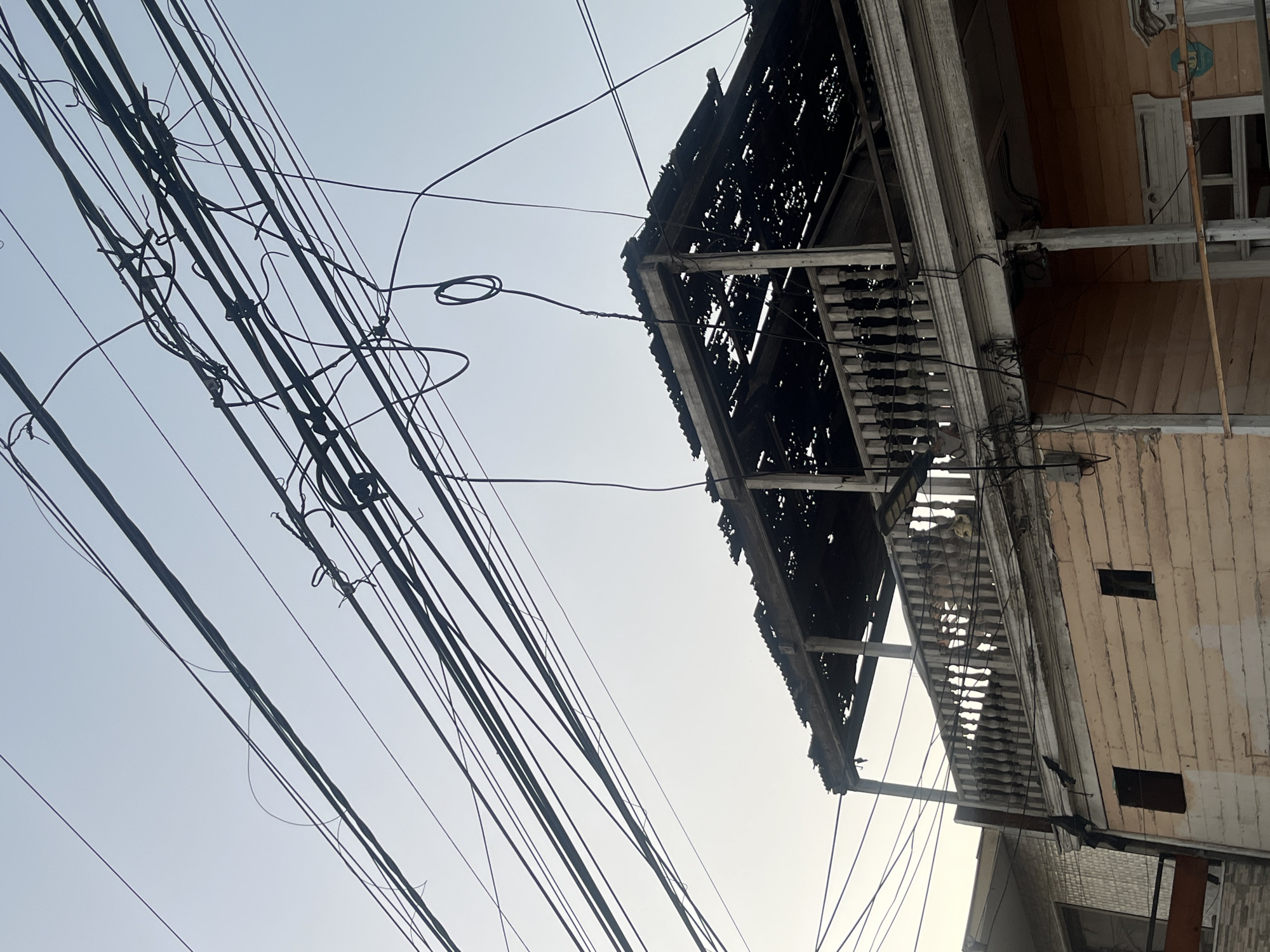 The image depicts a dilapidated building, possibly a house, in an urban environment. The building's facade is weathered and worn, with visible signs of decay. It features a balcony with deteriorated railings and a darkened, partially collapsed roof. Numerous electrical wires crisscross the sky in the foreground, adding to the urban setting. The sky is overcast and pale, suggesting a hazy or cloudy day. The photograph appears to have been taken from a low angle, emphasizing the building's height and the density of the overhead wires.