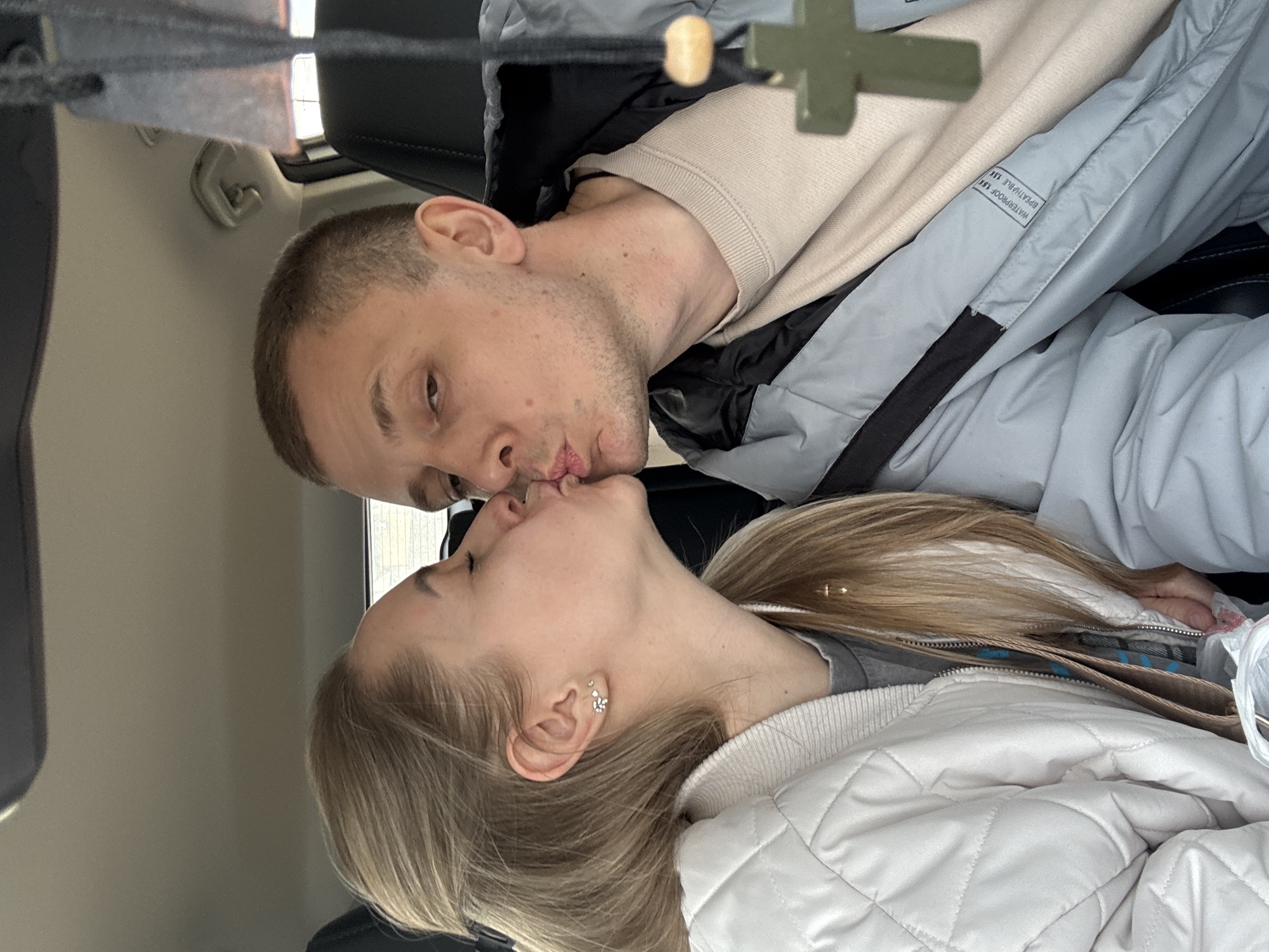 The image captures an intimate moment between a young man and woman, who are kissing inside a car in Chaikovsky, Russia. The man, with short dark hair and light stubble, is wearing a beige crewneck sweatshirt layered under a grey and black jacket. His eyes are partially visible, and his lips are pressed against the woman's. The woman has long blonde hair, which cascades over her shoulder, and multiple small, sparkling piercings in her right ear. She is wearing a grey top beneath a white quilted jacket.
A prominent detail in the upper part of the frame is a wooden cross pendant, hanging from above, suggesting it might be suspended from the car's rearview mirror or headliner. The setting appears to be the interior of a vehicle, with parts of the car's headliner and dark seat upholstery visible. On the sleeve of the man's grey jacket, specific text is visible: "WATERPROOF 10K" and "BREATHABLE 10K," indicating features of the garment. The overall mood conveyed is one of affection and closeness.