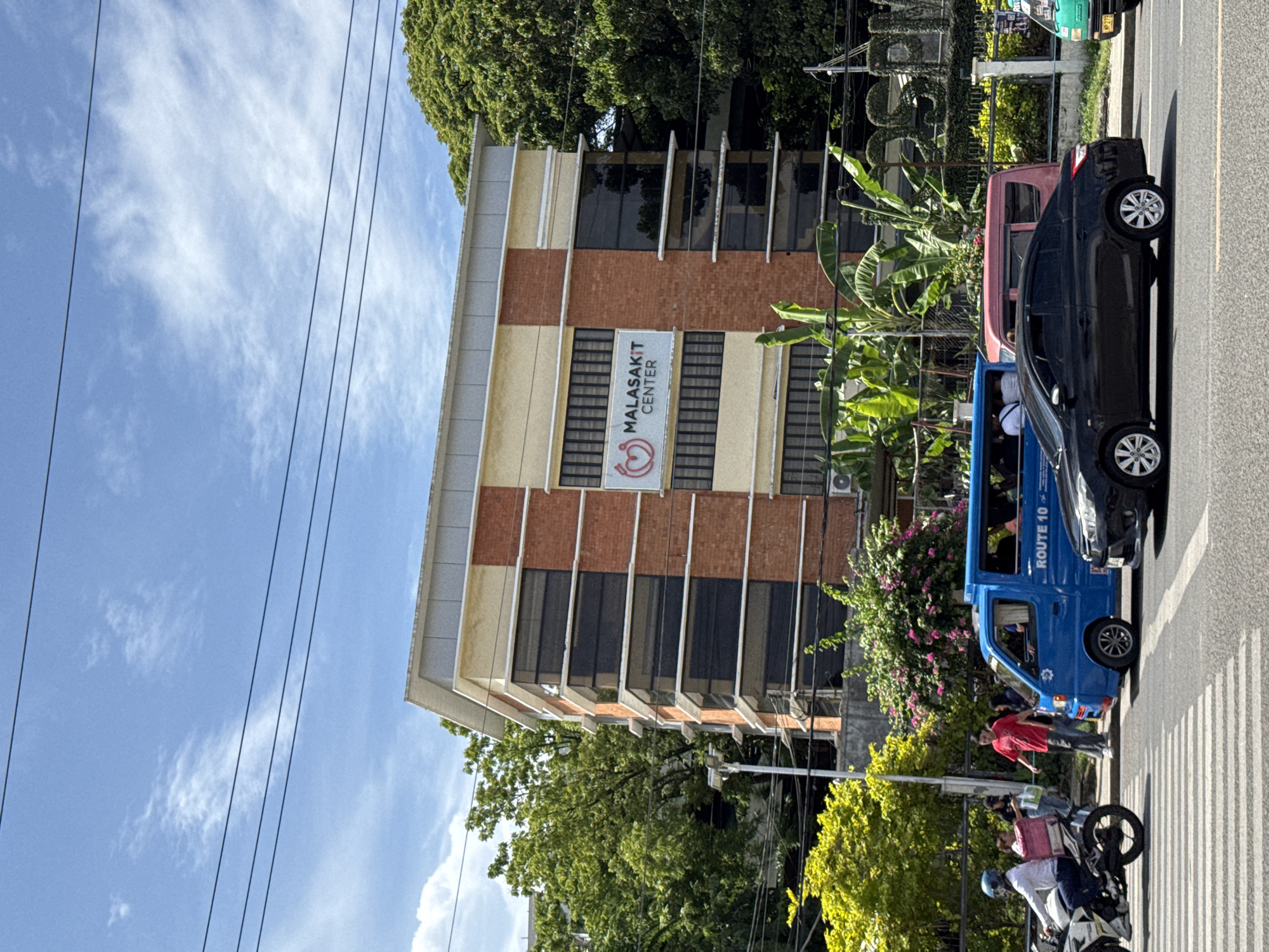 The image captures a vibrant street scene in Davao City, Philippines, under a clear blue sky with scattered white clouds. The bright sunlight suggests it is daytime, likely mid-morning or afternoon.
A multi-story building dominates the left side of the frame, featuring a light yellow facade and an upper section clad in orange-brown brickwork. Rectangular windows with horizontal elements are visible across its levels. A prominent white sign on the building clearly displays "MALASAKIT CENTER" in dark blue text, accompanied by a red heart-shaped logo. Lush green trees are visible behind and next to the building, including large, leafy canopies and banana plants.
On the right, several vehicles are parked or moving slowly along an asphalt road with visible pedestrian crossing markings. A black sedan, possibly a Toyota Vios, is prominent. Beside it is a blue jeepney-like public utility vehicle, which has "ROUTE 10" written on its side. A portion of a smaller pink/red vehicle is visible behind the blue one. Further down the street, a green vehicle, likely another public transport, is partially obscured. On the far right, near the pedestrian crossing, a white motorcycle is present, with a person wearing a blue helmet. Another person in a red shirt stands near the blue vehicle on the roadside. Overhead power lines crisscross the sky. The scene is bustling with typical urban activity.