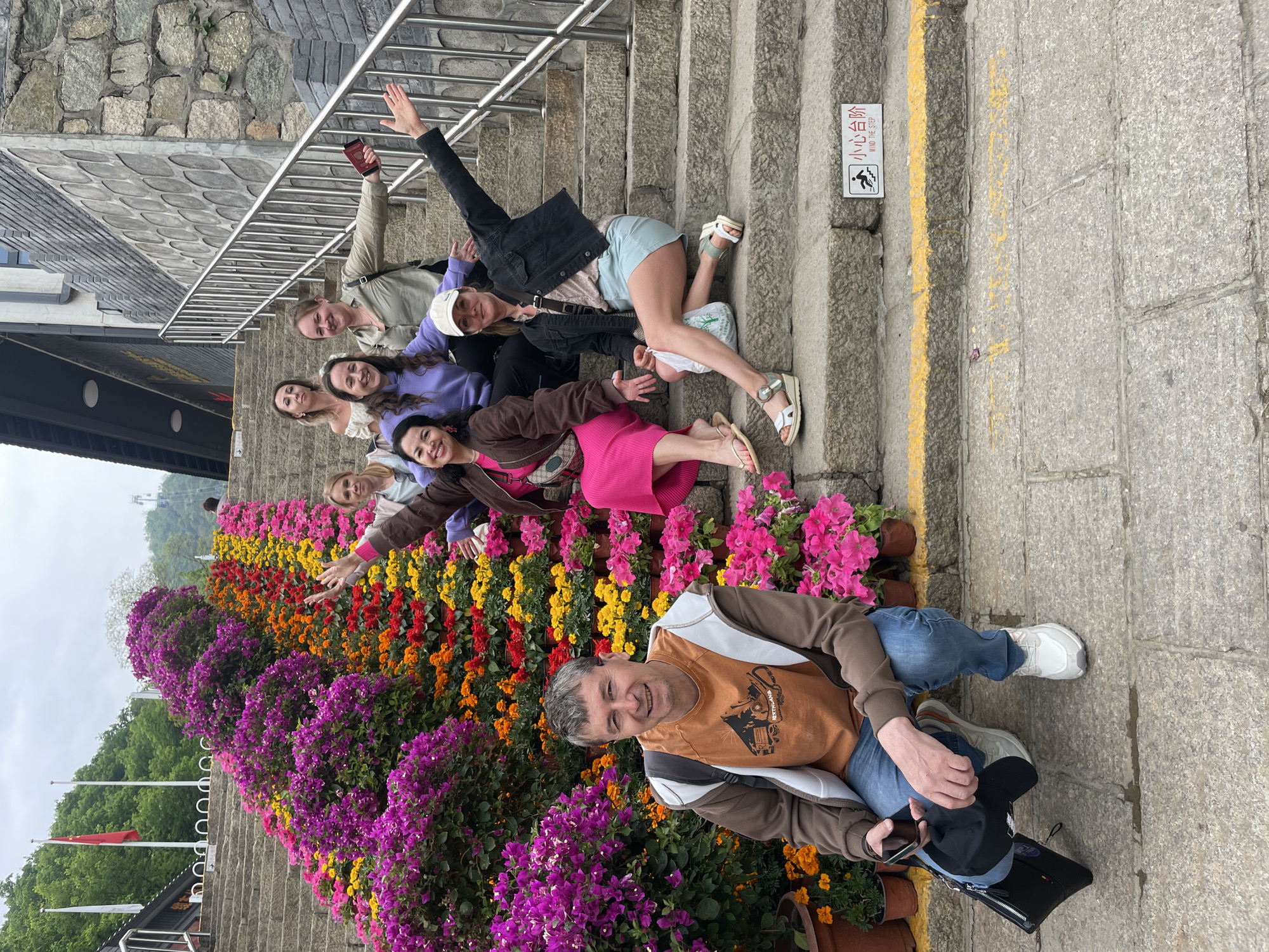 A group of eight people are posing on steps adorned with potted flowers. The individuals are positioned at varying levels of the staircase, with some seated and others leaning back or extending their arms in a playful manner. The steps are lined with flowers in shades of purple, orange, yellow, and pink.
The environment includes a metal handrail, a yellow painted line, and a sign in Chinese and English that reads "Mind the Step." The steps appear to be constructed from stone. Based on the flowers, stonework, and signage, the location is likely related to a tourist attraction or park within the city of Huairou District, China.