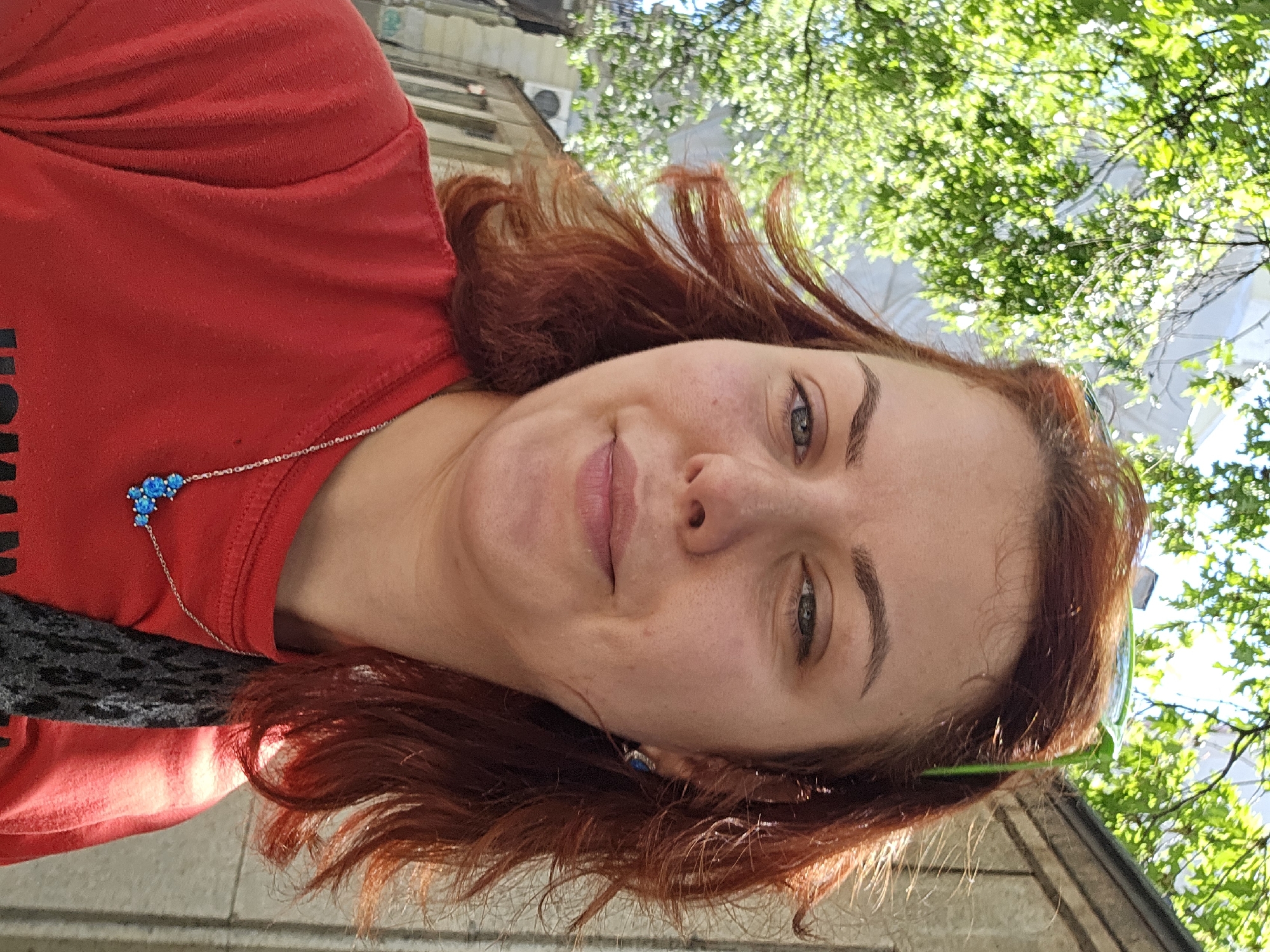 An outdoor, waist-up, selfie of a woman with auburn hair and fair skin is captured on a bright, sunny day. The woman is wearing a red t-shirt and a silver necklace with blue pendants. She has a slight smile and is looking directly at the camera. Her hair is windswept, with strands blowing across her face. In the background, there are lush green trees and a building with a textured wall. The lighting suggests it is daytime, with sunlight casting shadows and highlighting the woman's features. The environment appears to be a city street or park.