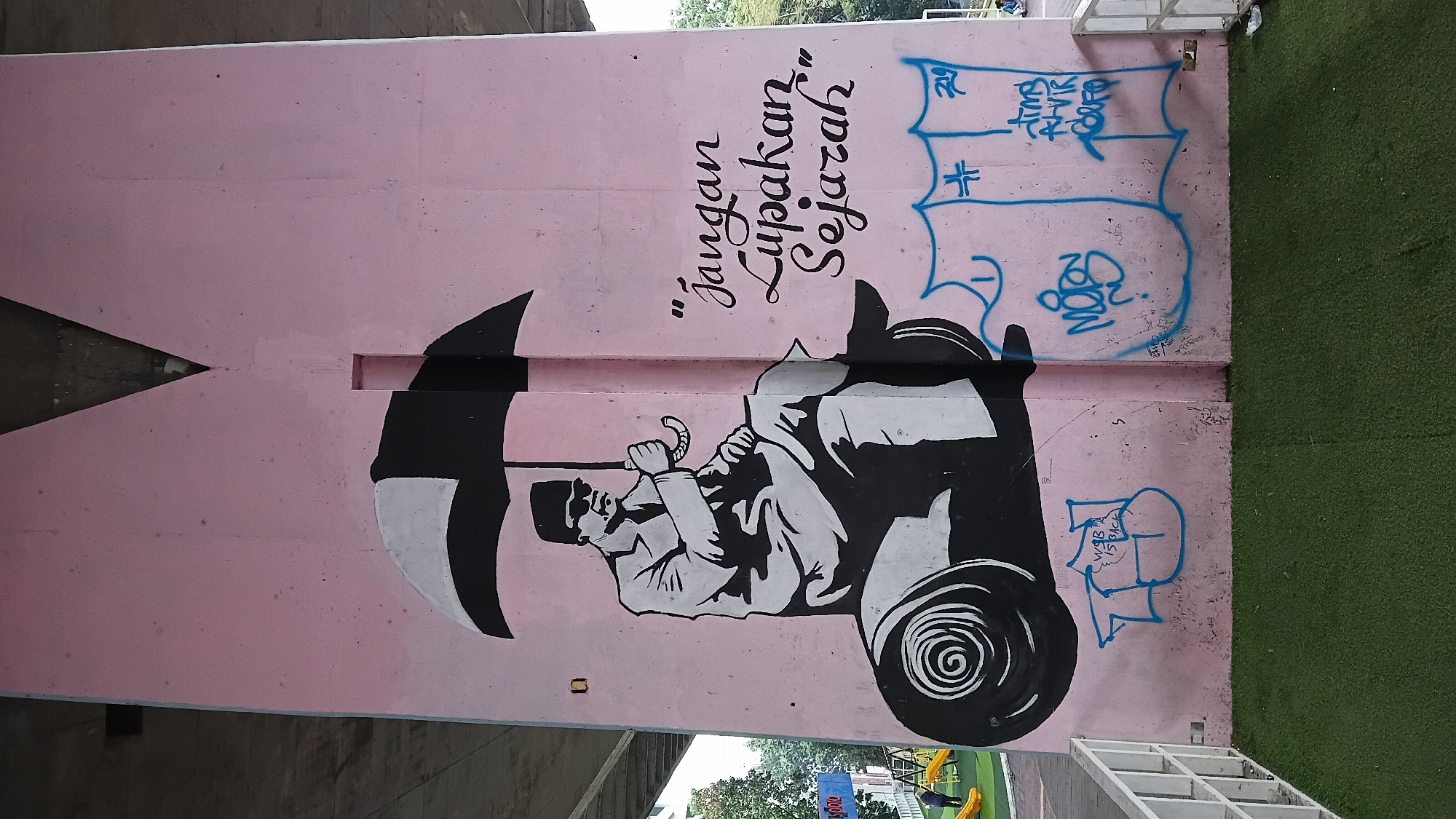 The image shows a mural painted on the side of a pink concrete pillar, likely part of an overpass or bridge structure, in Bandung City, Indonesia. The mural depicts a black and white stencil-style illustration of a man wearing a cap and sunglasses, seated on a large, rolled-up object that resembles a carpet or a large scroll. He is holding an open umbrella with black and white panels. To the right of the illustration, in black cursive script, are the words "Jangan Lupakan Sejarah," which translates to "Don't Forget History." Below this text, there are several tags or graffiti in blue spray paint, including what appears to be the symbol "#" and some illegible script. The pillar has a triangular architectural element at the top and a rectangular cutout in the middle. The base of the pillar is adjacent to a green artificial turf surface and a white metal railing. In the background, to the left, a playground structure with yellow slides and a person are visible. The environment suggests an outdoor urban setting, possibly a public space or park.