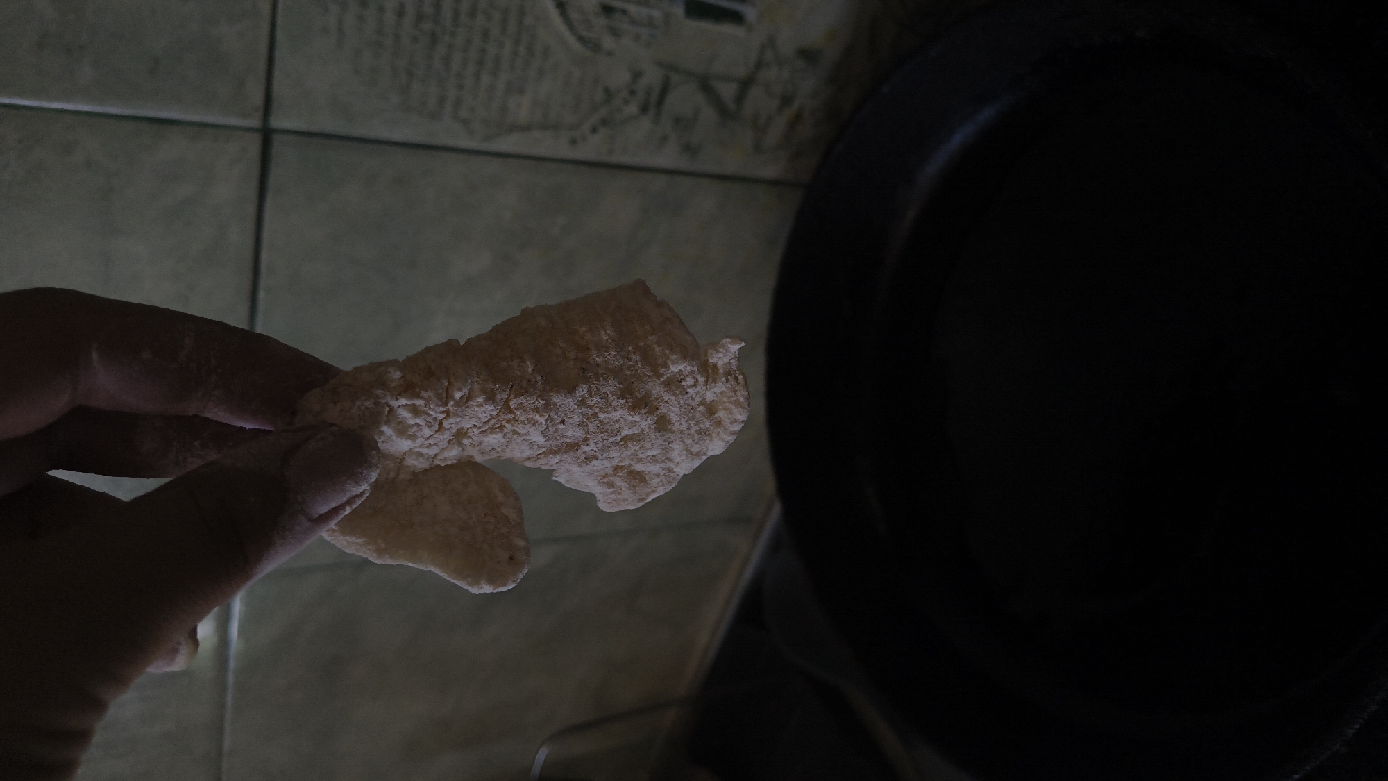 A close-up shot shows a person's hand holding two pieces of battered food over a dark frying pan. The food appears to be chicken tenders or fish sticks, coated in a thick, rough batter. The hand is dusted with flour, suggesting the food is being prepared for frying. The background consists of a tiled wall with a subtle pattern and what appears to be a partial view of a kitchen counter or appliance. The overall lighting is dim, with a spotlight effect on the food and hand. There is no information about the location in Vinnytsia, Ukraine, or any other city.