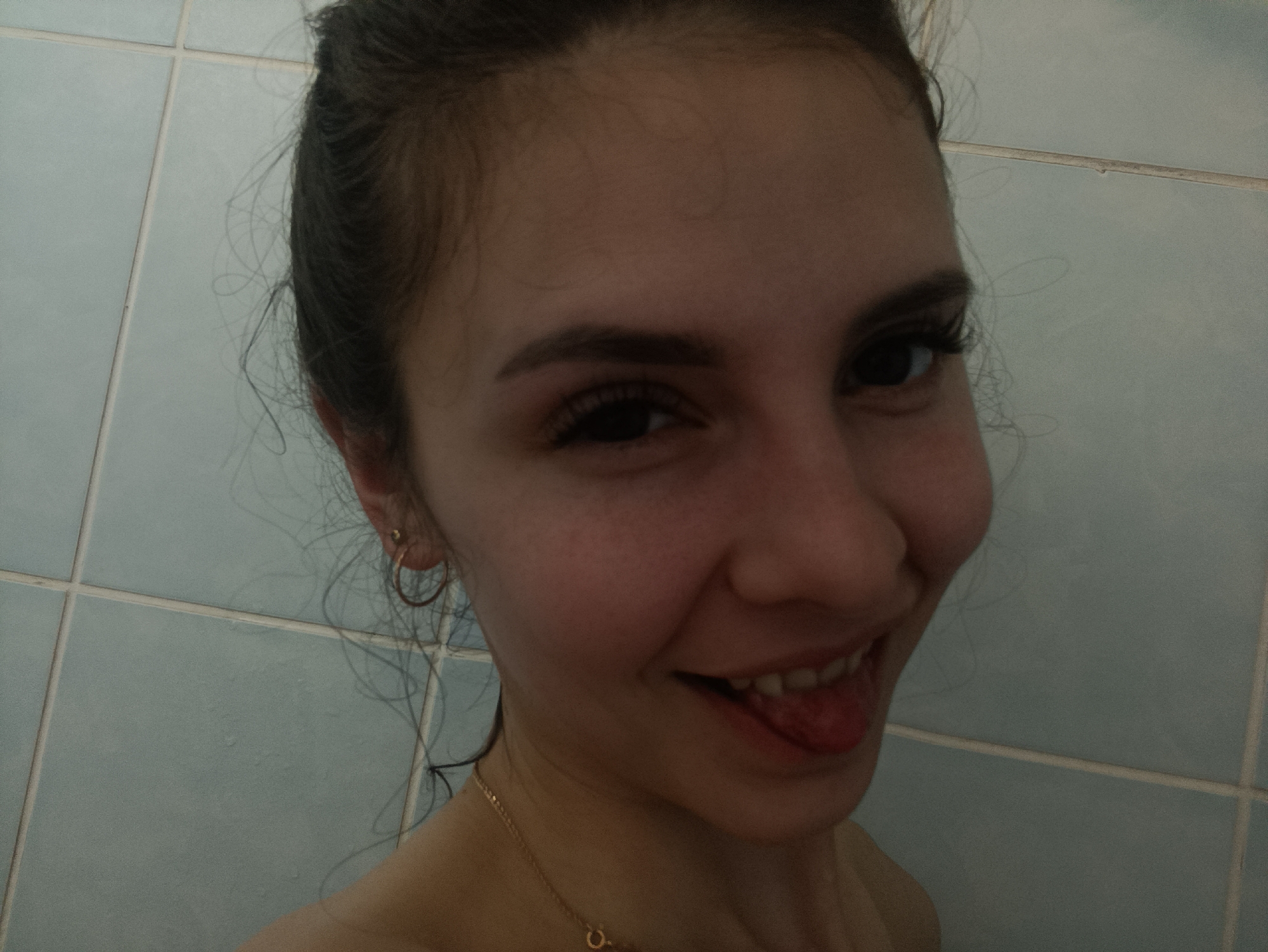 A young woman with dark hair pulled back into a bun is posing for a selfie in a bathroom with blue-gray tiled walls. She is smiling and sticking out her tongue. She is wearing gold hoop earrings and a gold necklace.