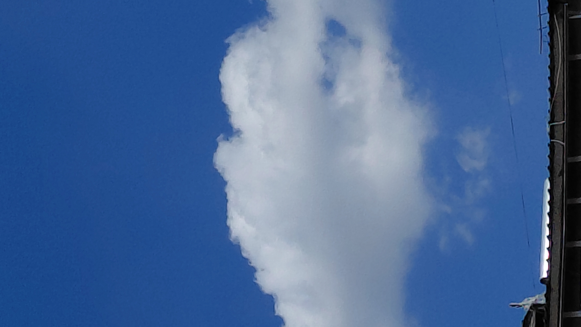 The image shows a bright blue sky with a large, fluffy white cloud spanning across the middle. The lower portion of the image shows a rooftop, likely made of dark corrugated material, with some metallic elements reflecting light. The scene is likely outdoors on a clear day. No people or specific landmarks are visible to determine the exact location within Abovyan, Armenia.