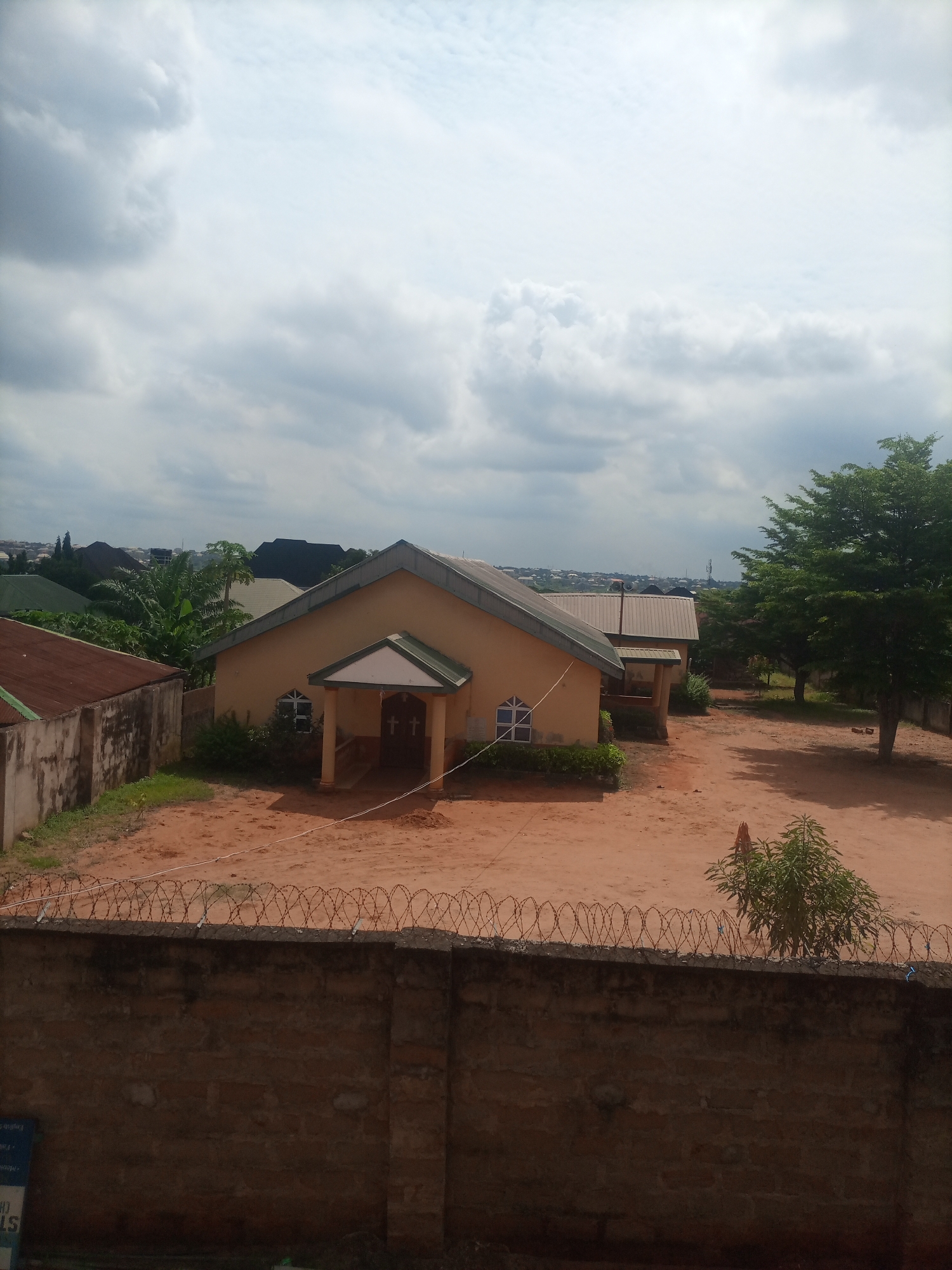 The image captures a view of a church building in Nkpor, Nigeria, under a cloudy sky. The church is a modest, single-story structure with a light yellow facade and a green-trimmed entranceway. Double doors with cross designs are visible at the entrance. The building is situated on a plot of red earth, with a scattering of green shrubs and bushes around it. To the left of the church, a smaller building with a corrugated metal roof is visible. In the background, a dense cluster of trees and other buildings suggest a developed urban or semi-urban environment. The foreground is dominated by a brick wall topped with barbed wire, indicating a property boundary. The overall scene conveys a sense of a functional, everyday religious institution within a Nigerian town.