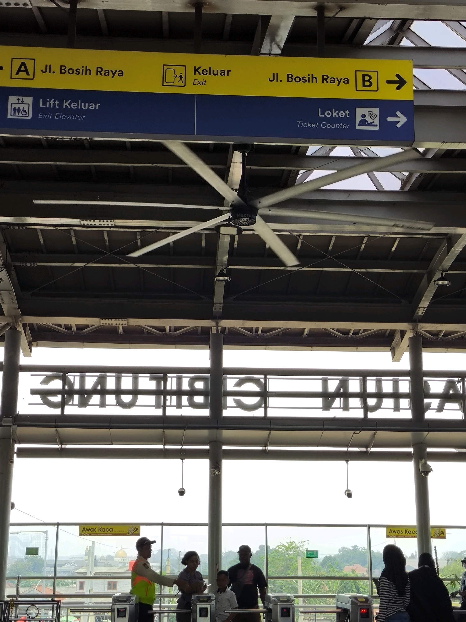 This image captures a scene at a transportation hub in Mekarsari, Indonesia, likely a train or bus station, indicated by the signage and fare gates. In the foreground, a large industrial fan is suspended from the ceiling, its blades slightly blurred due to motion. Below the fan, directional signs are prominently displayed.
The top sign, in yellow and blue, directs to "A Jl. Bosih Raya" and "Keluar" (Exit) on the left side, and "Jl. Bosih Raya B" with an arrow pointing to the right on the right side. The lower sign, in blue, indicates "Lift Keluar" (Exit Elevator) with accessibility symbols on the left, and "Loket" (Ticket Counter) with a person icon and an arrow on the right.
Further down, through a large windowed area, several individuals are visible. A uniformed security guard in a fluorescent vest is interacting with a woman and a child, who are standing near a fare gate. To the right, another woman wearing a hijab is looking at her phone, also near the fare gates. In the background, a cityscape with buildings and lush greenery is visible, including a prominent golden-domed building, possibly a mosque. Another sign that reads "Awas Kaca" (Beware of Glass) is also visible near the fare gates. The overall environment suggests an indoor waiting area with an outdoor view.