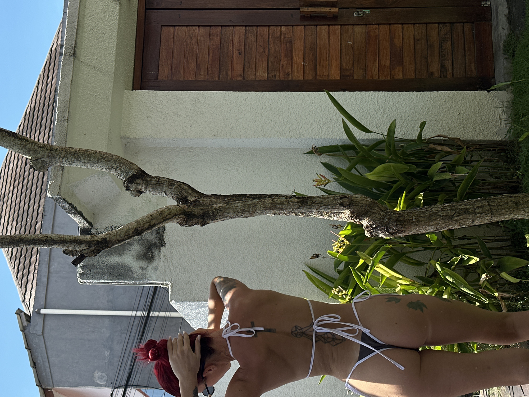 The image captures a person with red hair standing outdoors in Dalung, Indonesia. The individual is wearing a bikini. The surroundings include foliage and what appears to be a building with a wooden door. A tree branch extends across the frame, partially obscuring the building. The sky is visible in the background. Electrical wires run in the background.