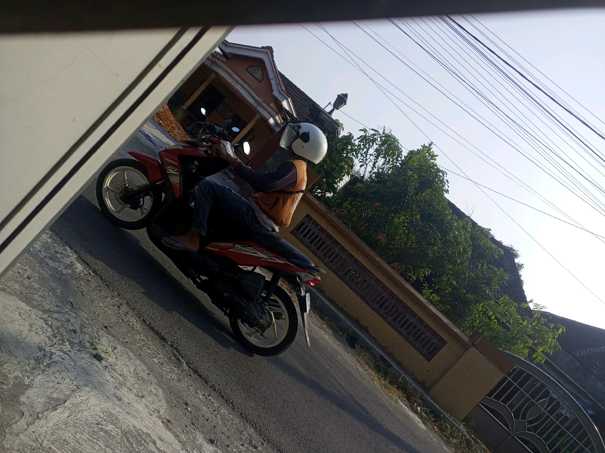 The image shows a person on a red Honda motorcycle parked on an asphalt road in Ngasem, Indonesia. The person is wearing a white helmet and an orange vest over a dark long-sleeved shirt and dark pants. The motorcycle is angled towards the left of the frame, with its front wheel turned. To the left of the motorcycle, a portion of a building is visible, with a white architectural element in the foreground. Behind the motorcycle, a yellowish-brown wall with a decorative metal gate runs along the property line. Lush green trees and the roof of a house are visible beyond the wall. The sky is clear and light blue, with numerous electrical wires strung across the upper portion of the image. The lighting suggests it is daytime, with the sun casting shadows on the road and the side of the building.