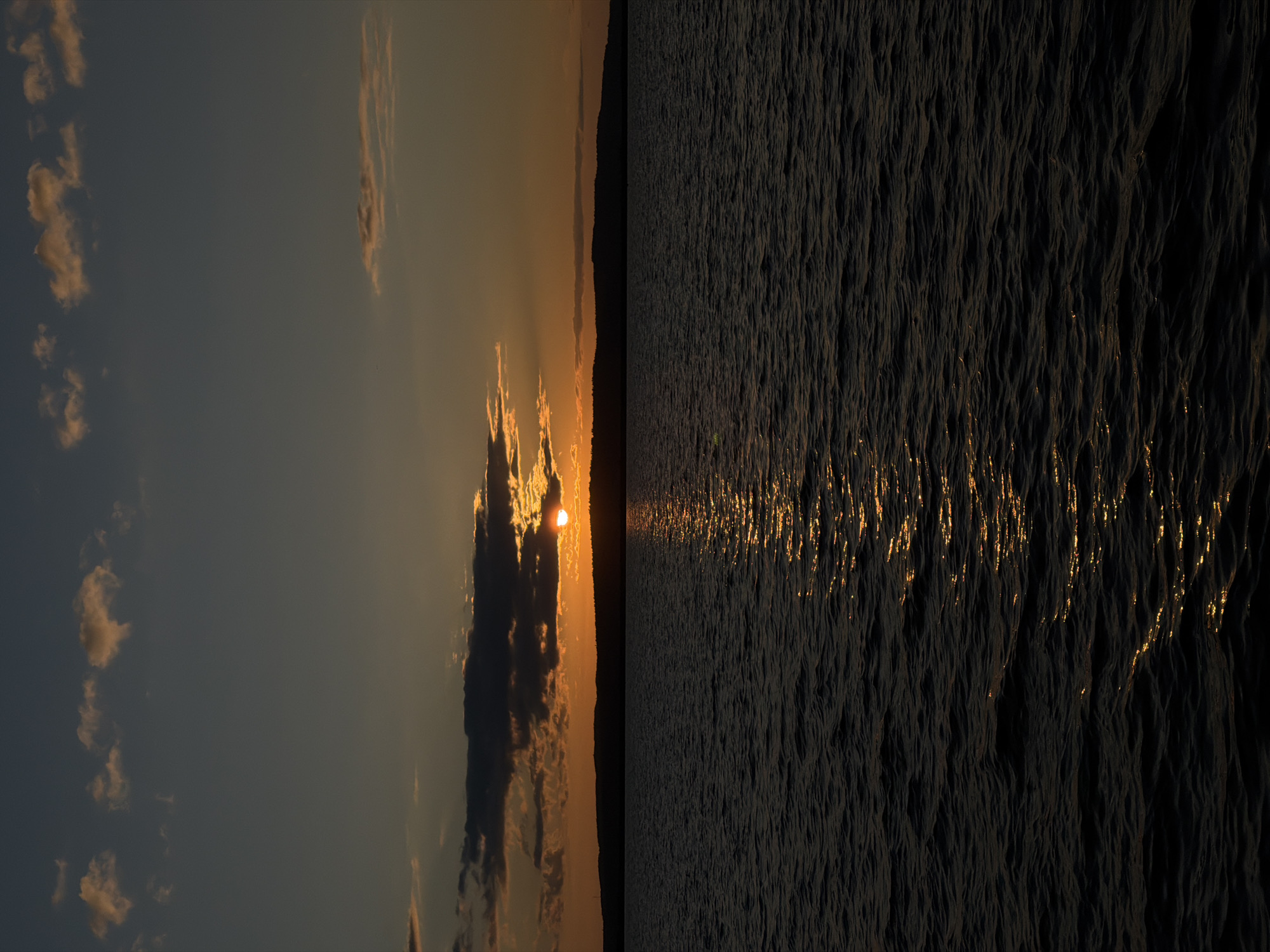 A vertical, medium shot captures a tranquil sunset over a body of water, likely the Kama River in Chaikovsky, Russia. The sun, partially obscured by dark clouds, casts a warm orange glow on the horizon, illuminating a band of lighter clouds above. The dark silhouette of a distant, forested shoreline stretches across the middle ground. The foreground is dominated by the dark, rippling surface of the water, reflecting the golden light of the setting sun in a shimmering path that leads the eye toward the horizon. The sky transitions from a deep twilight blue at the top to a warm orange hue near the horizon. There are no people or man-made structures visible in the frame, creating a sense of serene, natural beauty.
