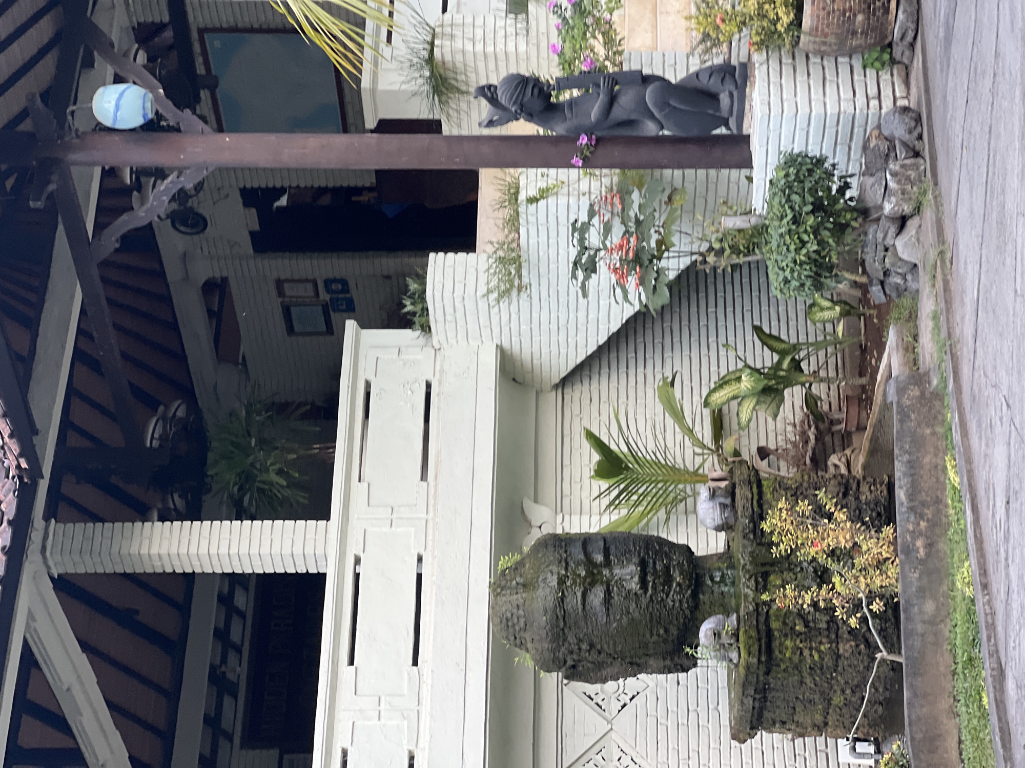 The image depicts the exterior of a building with white brick walls and a dark tiled roof. A large stone Buddha head sculpture, covered in moss and small green plants, is positioned in the foreground, partially obscured by a concrete structure. To the right of the Buddha head, a dark-colored statue of a seated figure holding a long, thin object is visible. The building features a white balcony with decorative elements and a set of white brick stairs leading upwards. Several potted plants and greenery are scattered throughout the scene. A blue and white glass lantern hangs from a wooden beam above the entrance. The environment appears to be an outdoor setting, possibly a courtyard or garden area, in Abang, Indonesia.