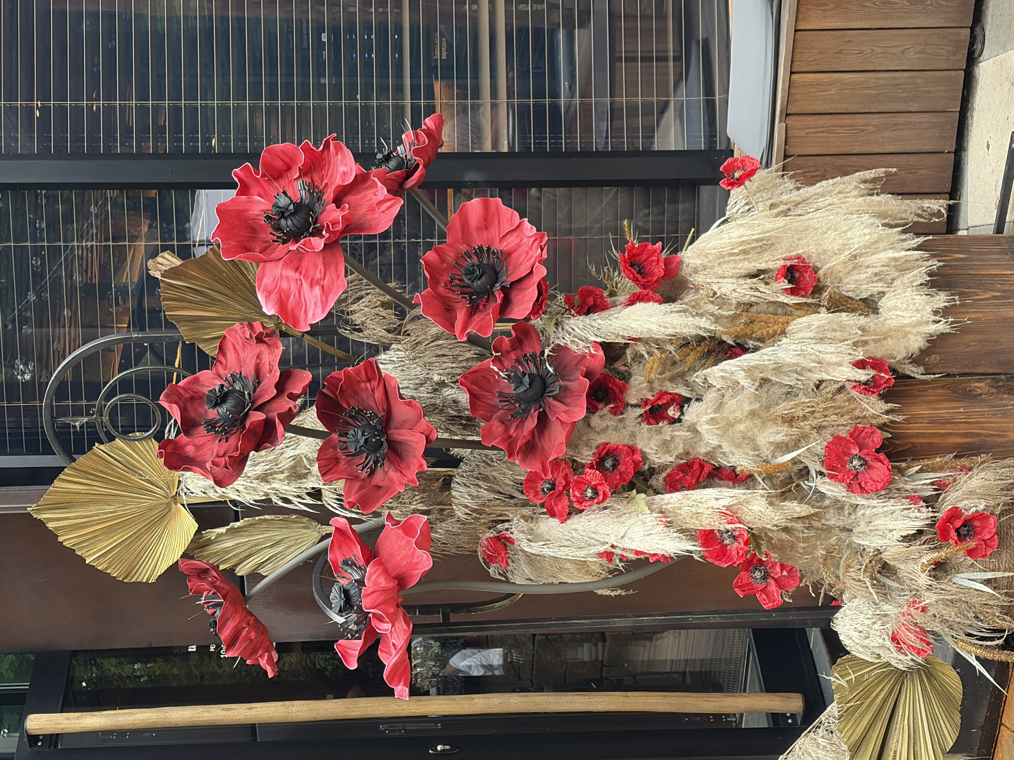 An outdoor floral arrangement is prominently displayed in front of a building. The arrangement features large, red artificial poppies with black centers, interspersed with dried beige grasses and a few golden fan-shaped leaves. The poppies vary in size, with some significantly larger than others. The arrangement is housed in a dark wooden planter.
Behind the arrangement, the building has large windows with horizontal blinds, reflecting the environment outside. A vertical wooden beam is visible to the left of the arrangement. The ground in front of the planter appears to be paved. In the reflection of the window, a person wearing a white shirt is visible.
While the image itself does not provide definitive evidence of the specific location within Khimki, Russia, similar floral displays are often found outside establishments such as cafes, restaurants, or shops in urban settings. The style of the building and the surrounding paved area suggest a commercial or public space.