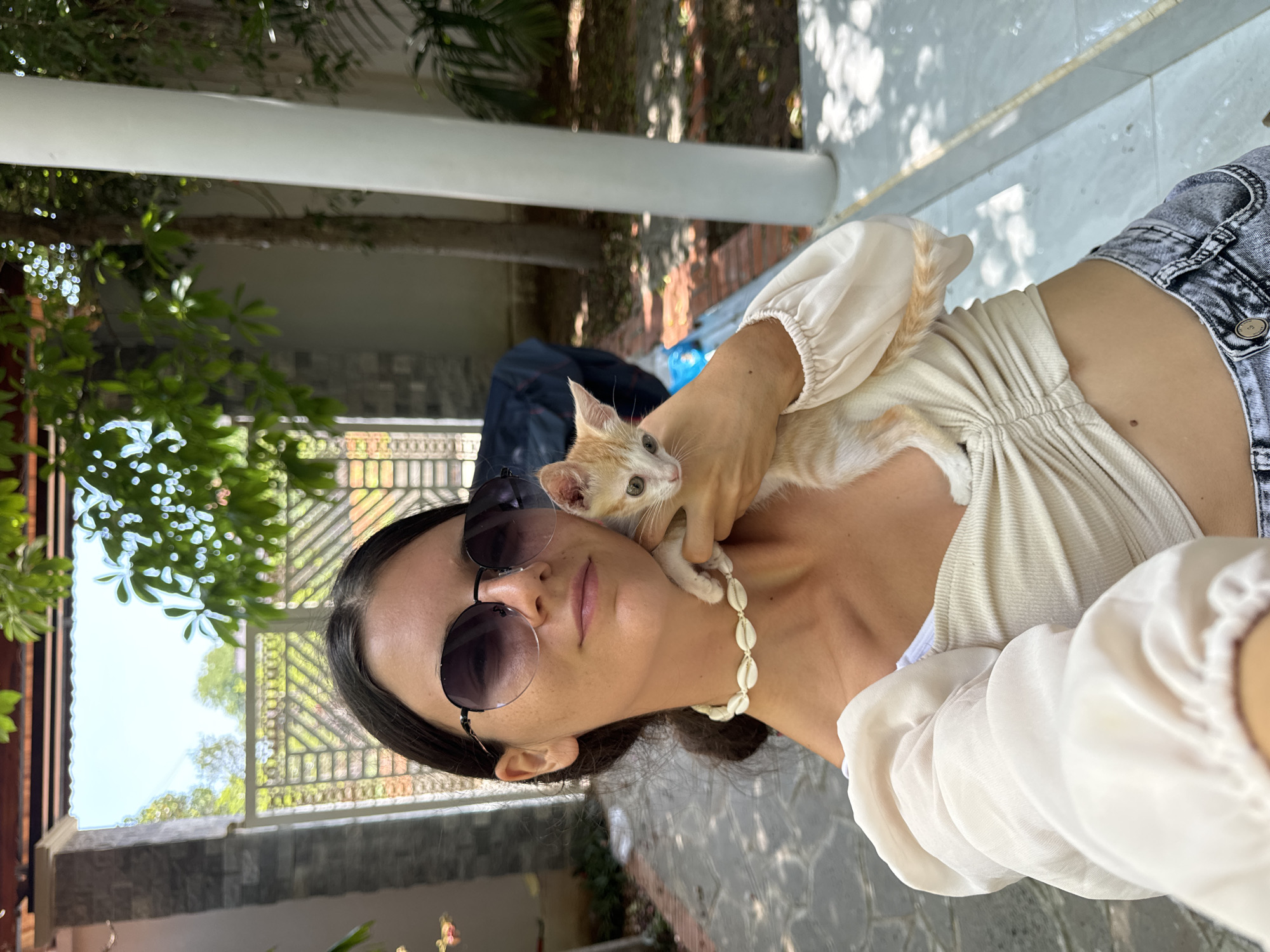A young woman with dark hair and sunglasses takes a selfie holding a small, light-colored kitten. She is wearing a cream-colored, ruched top and denim shorts. She has a shell necklace around her neck. The woman and kitten are outdoors, in what appears to be a courtyard or garden area with tiled flooring and brick pathways. In the background, there is a white metal gate, stone pillars, and lush green foliage, suggesting a tropical environment. The sunlight is bright, casting shadows on the ground. This scene is likely located in Mũi Né, Vietnam, given the architectural elements and vegetation typical of the region.
