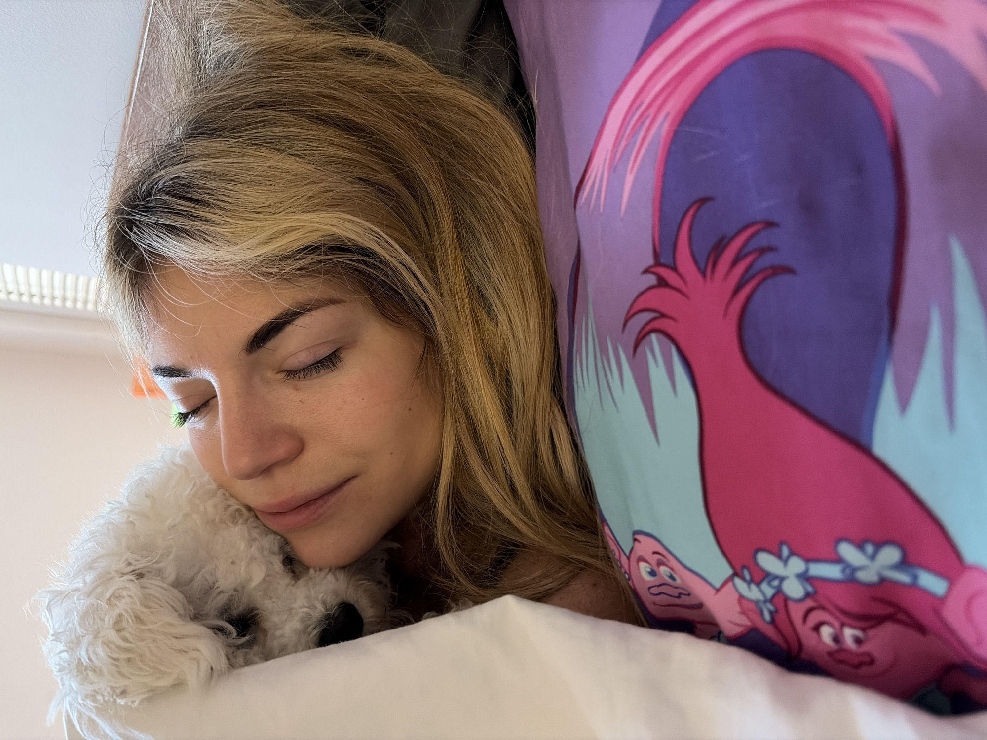 A woman with long, blonde hair rests her head on a small, white dog with curly fur, presumably in a bed with white linens. A colorful pillowcase featuring a cartoon troll is visible to the right of the woman. Light streams in through a partially visible window. The location of this photo is not evident within the scene.