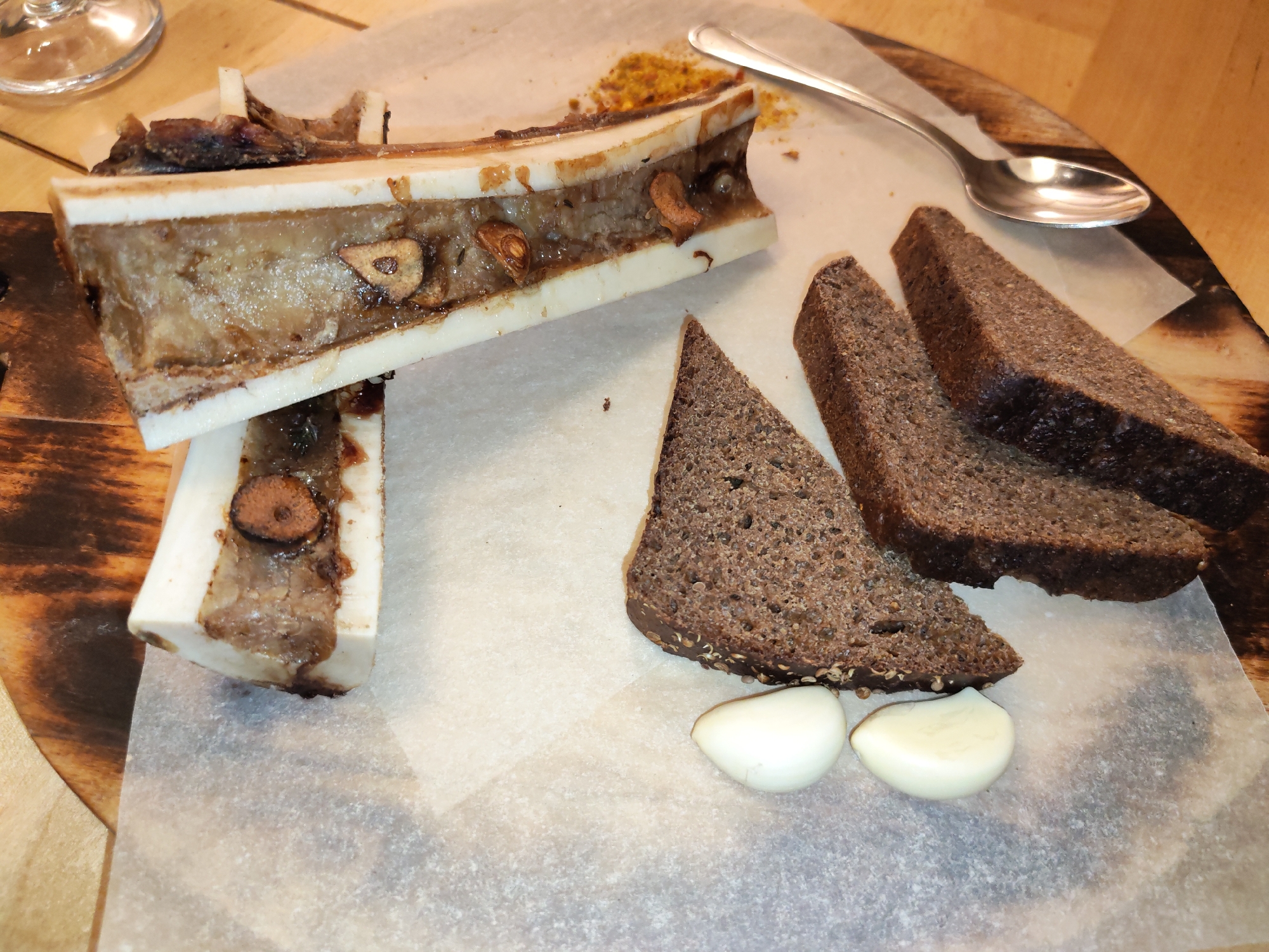 The image presents a culinary arrangement featuring bone marrow. Two bone segments, split lengthwise, showcase the cooked marrow infused with garlic cloves. Adjacent to the bones are slices of dark rye bread and two cloves of raw garlic. A spoon rests nearby, suggesting the marrow is to be extracted and spread onto the bread. The presentation is on a wooden board covered with parchment paper. The environment is a restaurant, likely located in Izhevsk, Russia, indicated by the food being a Russian cuisine. No people are visible in the image.