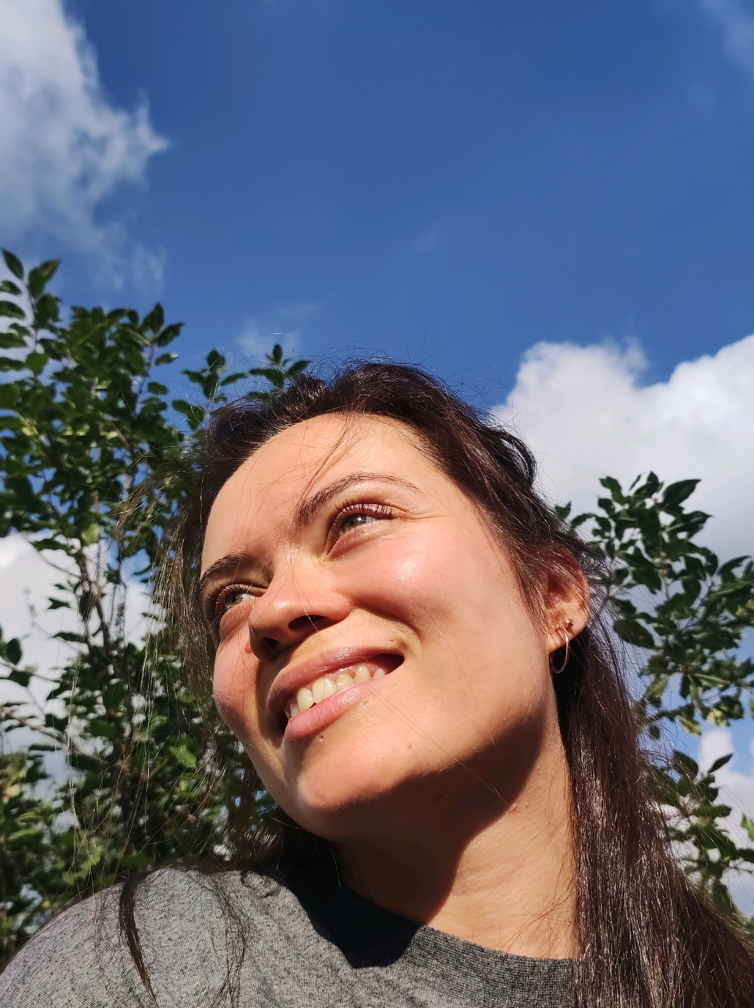 A close-up, low-angle shot captures a young woman smiling and looking upwards against a bright blue sky with scattered clouds and green foliage. The woman has dark brown hair, and her face is illuminated by sunlight, casting a warm glow. She is wearing a grey t-shirt. The background consists of a tree with green leaves and the sky. The image does not contain any information about the city of Фоки, Russia.