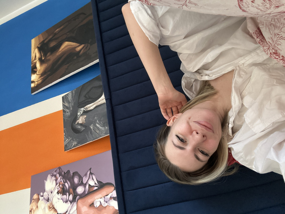 An indoor scene in the city of Khimki, Russia, depicts a woman resting on her left side in the foreground, facing the viewer. She has light-colored hair and a slight smile, with her left hand positioned behind her left ear. She is wearing a light-colored, long-sleeved collared shirt and patterned lower garments with a reddish-brown, intricate design. The woman is positioned against a dark blue, vertically tufted headboard or sofa back. Above this padded surface, leaning against a wall painted with vertical stripes of white, orange, and blue, are three artworks. From left to right: a canvas featuring a hand holding light-colored floral blossoms on a lavender background; a central darker canvas depicting a figure with dark hair and a pale complexion looking downwards, against a textured grey background; and a rightmost canvas displaying an abstract or stylized human form in brown and beige tones.
