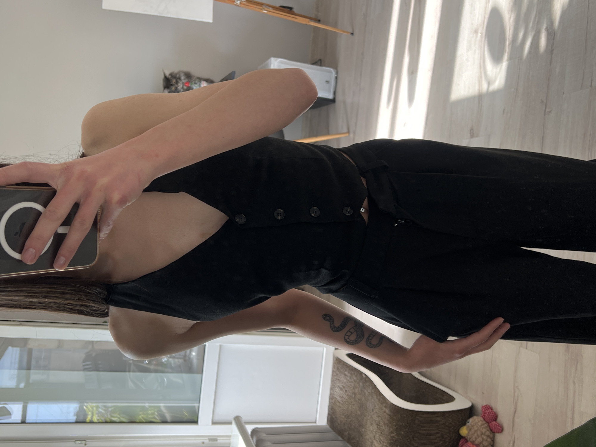 A person is taking a selfie indoors while wearing a black, button-up vest and black pants. The person has a snake tattoo on their forearm. In the background, there is a cat, a white object, and light coming through a window creating shadows on the floor. A cat tree is also visible, along with a small toy. There's no verifiable information to indicate that the image was taken in Ternopil, Ukraine.