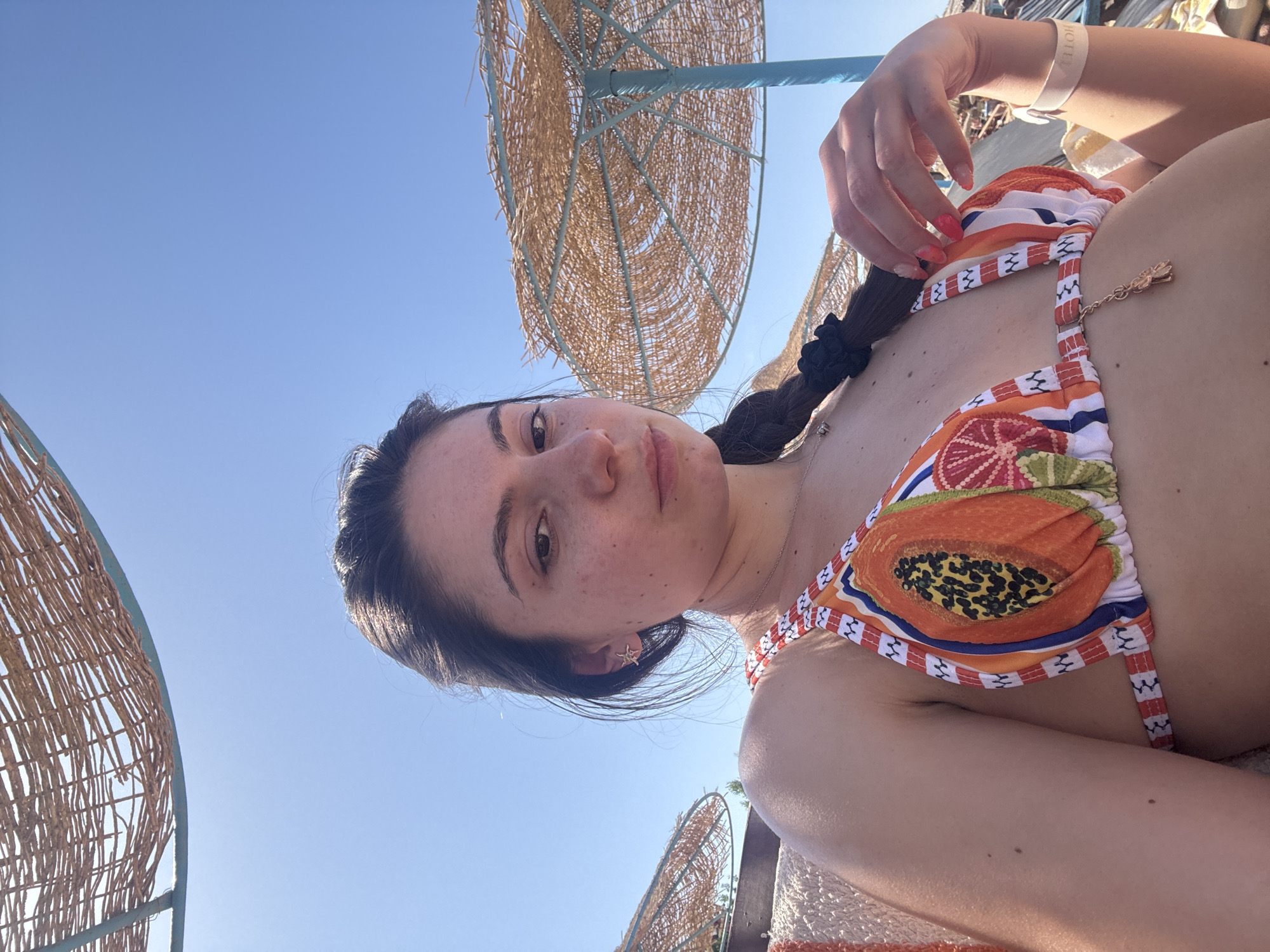A woman with fair skin and dark hair in a braid is lying down, facing the camera. She is wearing a colorful bikini top patterned with fruit designs. Her left hand is raised, holding a strand of her hair. She has a small gold charm hanging from her bikini top. A white wristband is visible on her right wrist. The scene is outdoors under a clear blue sky, and woven parasols are in the background. The location appears to be a beach, and the city and region are Unknown.