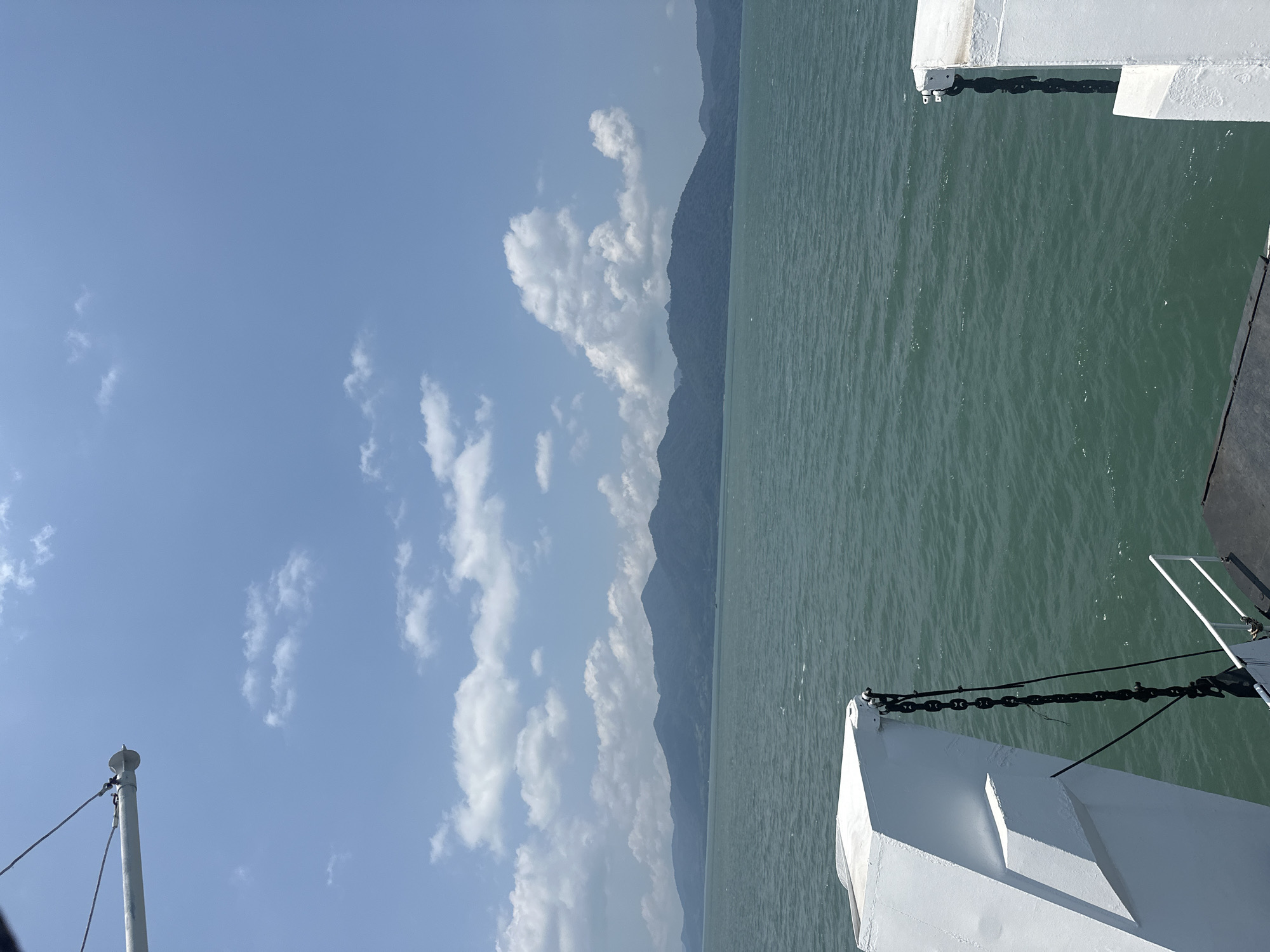 View from a ferry in Laem Ngop District, Thailand. The scene shows the ocean, mountains in the distance, a blue sky with white clouds, and parts of the white ferry in the foreground, including chains and a mast.