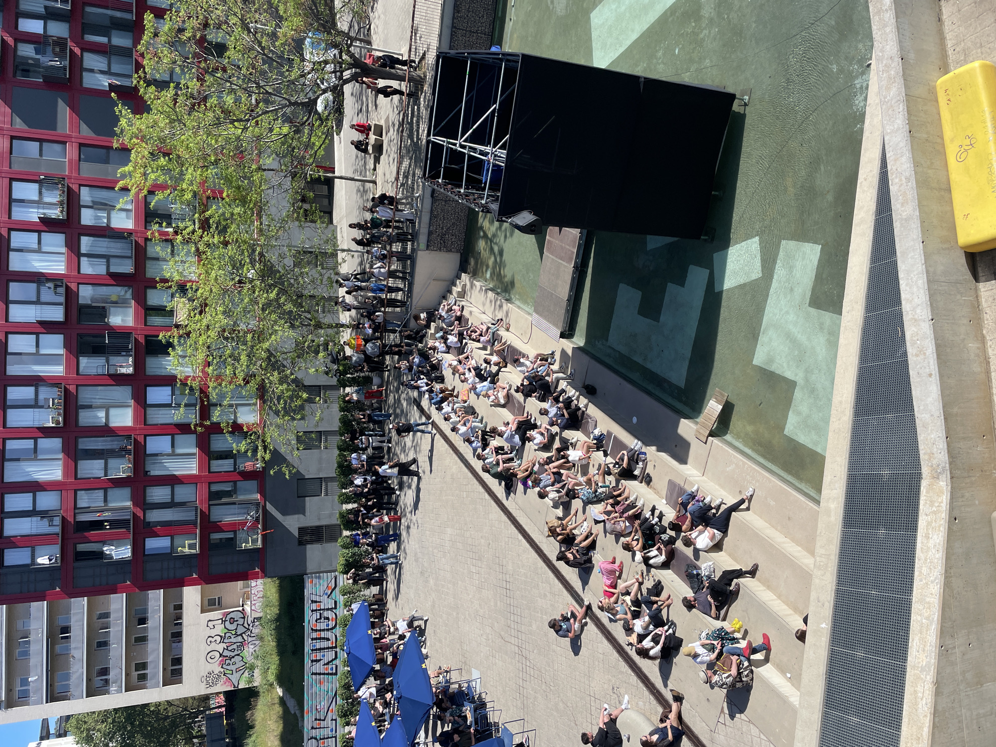 The image captures a high-angle view of an outdoor event in Barcelona, Spain. A crowd of people is seated on tiered concrete steps facing a stage structure partially submerged in a shallow pool. The stage is black, constructed with metal scaffolding. The pool has geometric patterns painted on the bottom. The crowd is primarily young adults.
To the left of the steps is a building with red frames around the windows and another next to it with graffiti on the walls. Several people are milling in the area behind the seated audience. To the left, blue umbrellas cover café tables. The overall lighting suggests a sunny day.