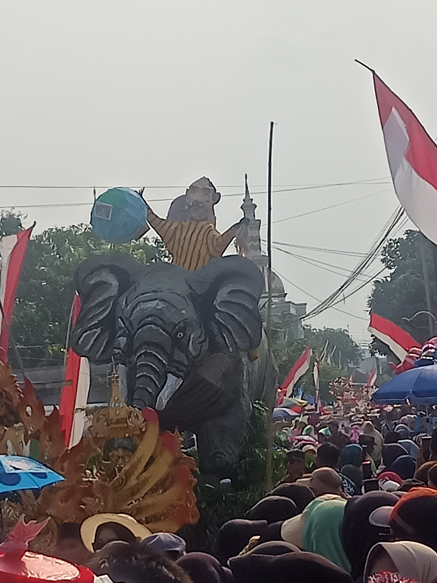 This is a photograph taken in Gemolong, Indonesia, on a day with overcast skies. The foreground is dominated by a large, dark gray, cartoonish effigy of an elephant, its trunk raised. A figure wearing a yellow and brown striped shirt and a headdress is seated on the elephant's head, holding a blue and green globe. Behind the elephant, a crowd of people are gathered, some holding red and white Indonesian flags. In the background, a mosque with a dome and minaret can be seen, along with trees and electrical wires. The overall scene suggests a parade or a public gathering.