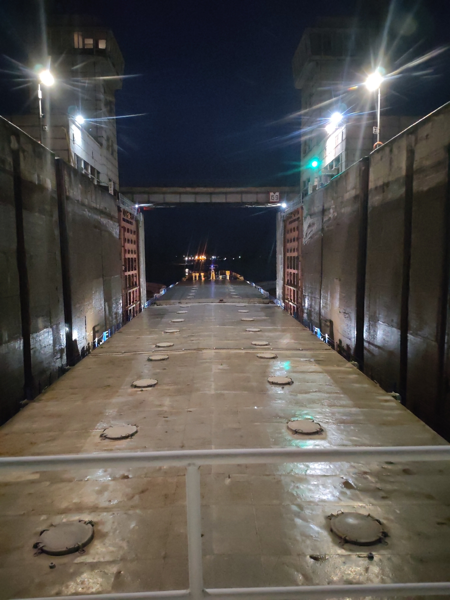 The image depicts the interior of a lock at night, with high concrete walls on either side. Overhead are structures, including a bridge with the number "159" visible. Lights illuminate the walls and the walkway below, where a series of round manhole covers are visible. A white railing is in the foreground. Beyond the lock, other lights are seen in the distance.