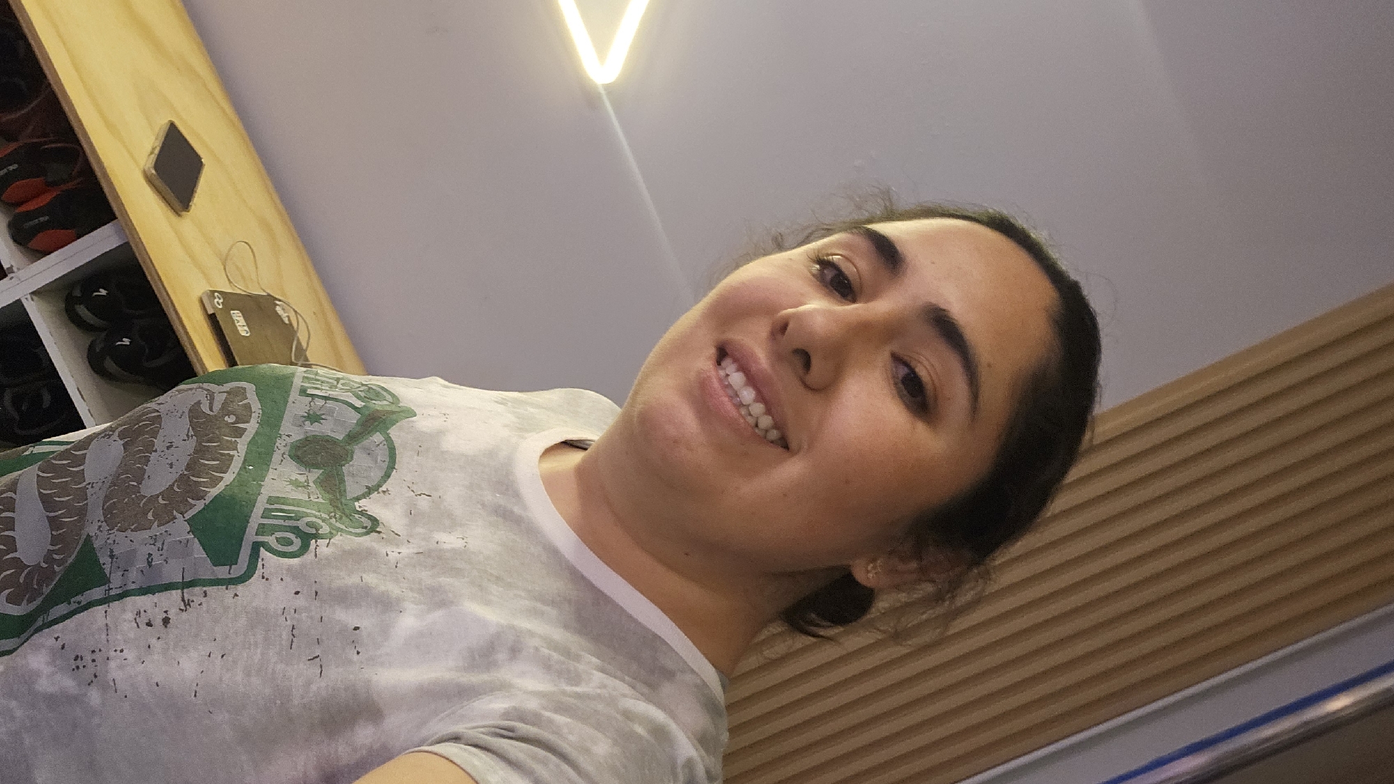 A selfie, taken at an upward angle, depicts a woman with dark hair and a light complexion. She is smiling and wearing a light grey t-shirt with a green and white design that includes a snake. The background includes a ceiling with a triangular light fixture, a wall with horizontal wood paneling, and a shelf with shoes and other items. Based on the emblem on the t-shirt, it is possible that this image was taken in Mexico, specifically in Puebla.