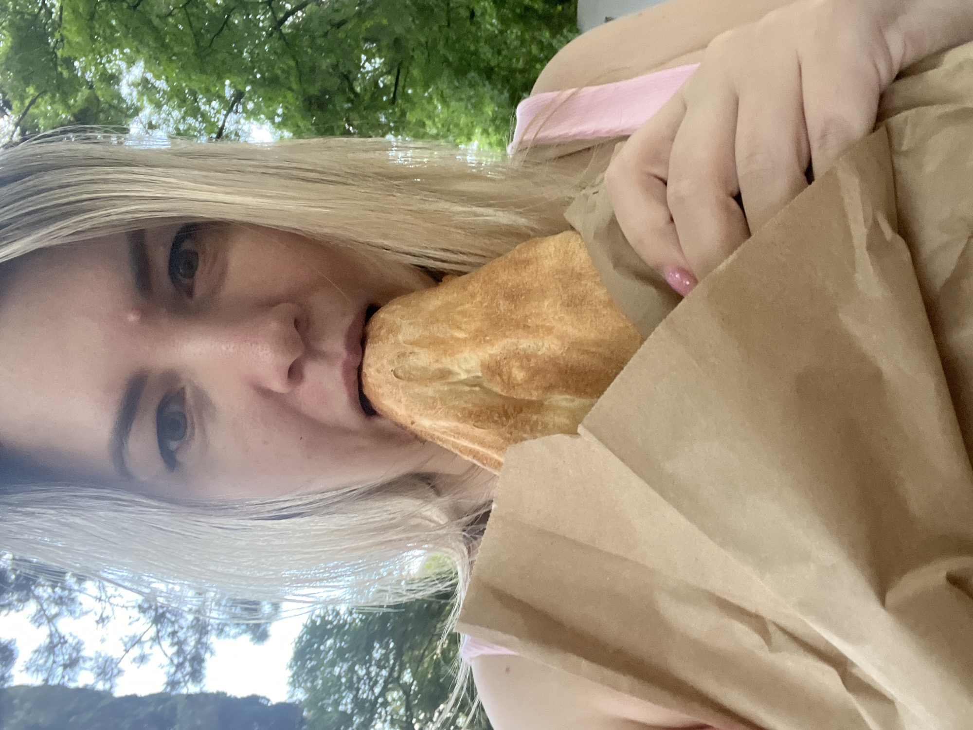 A blonde woman is taking a bite out of a pastry from a brown paper bag. She is wearing a pink top. The background shows green trees and a blue sky, suggesting an outdoor setting in Sochi, Russia.
