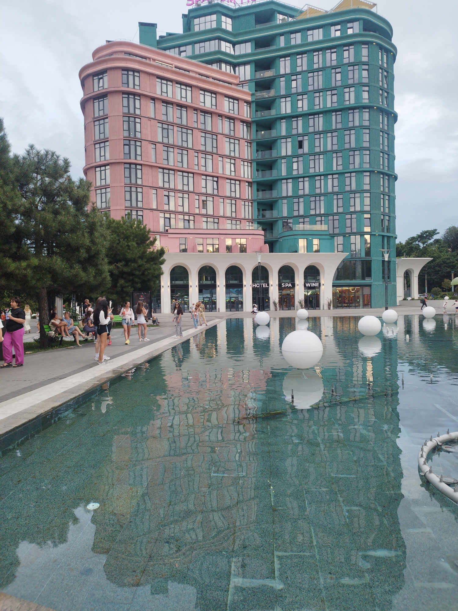 The image depicts a contemporary architectural complex situated in the city of Odesa, Ukraine. Dominating the scene are two adjacent buildings, one clad in a soft pink facade and the other in a teal hue. Both structures feature numerous windows and balconies. In the foreground, a water feature, possibly a fountain or reflecting pool, occupies the lower half of the image. Large, white spherical objects are partially submerged in the water. The water's surface reflects the buildings and the sky, creating a sense of depth and visual interest. Several people are visible in the background, strolling along a paved walkway adjacent to the water feature. Trees line the walkway, providing a touch of nature to the urban setting. The overall atmosphere appears to be one of a modern public space, possibly a plaza or promenade.