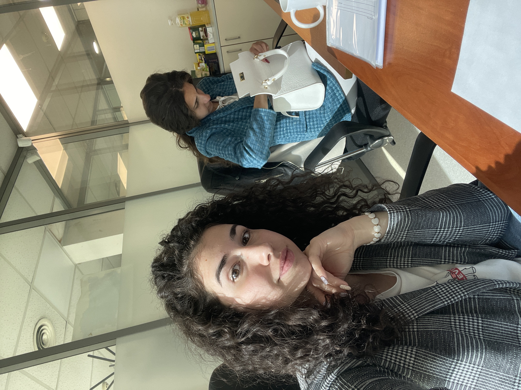 Two women are in an office-like setting. The woman in the foreground is sitting in a chair wearing a gray patterned blazer and has curly brown hair. She is holding her chin with her hand and looking directly at the camera. The second woman is sitting in a chair behind her and to the left, wearing a blue plaid jacket and looking at a white handbag she is holding. There is a wooden desk to the right with a cup and papers on it. Behind the second woman is a shelf with various bottles and containers on it. The room has large windows that suggest it is inside a building. The location is unspecified, but given the request, it is presumed to be within the city of Sofia, Bulgaria.