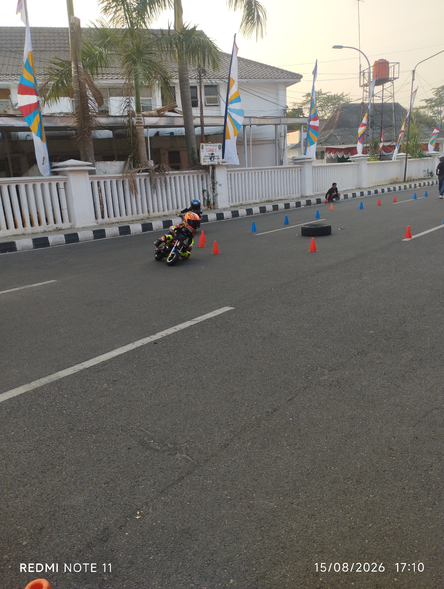 An outdoor, low-angle shot captures a child riding a miniature motorcycle on a paved road. The child, wearing a helmet and protective gear, leans into a turn, navigating around orange traffic cones. The road is marked with white lines and bordered by a white fence with black accents. Palm trees and a building with flags are visible in the background. The overall scene suggests a race or practice event. The image also features a timestamp in the lower right corner reading "15/08/2026 17:10" and "REDMI NOTE 11" on the lower left.