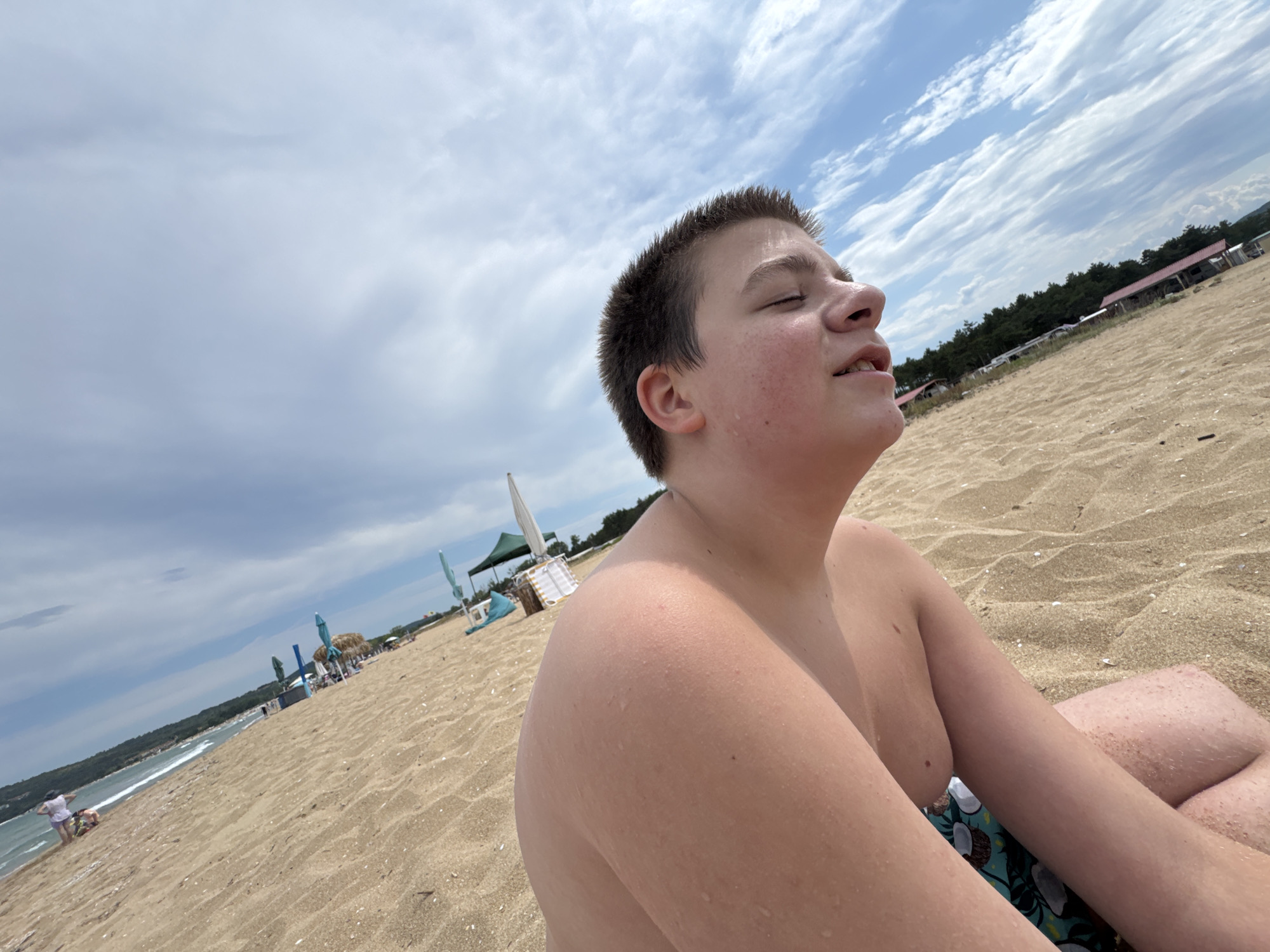 A young boy sits on a sandy beach in Sozopol, Bulgaria, with his eyes closed and face tilted upwards, seemingly enjoying the sun or a gentle breeze. The sky above is partly cloudy, with a mix of blue patches and white clouds. In the background, a line of trees and buildings can be seen along the coastline, indicating a developed area. Beach umbrellas and other beach equipment are scattered along the sand, suggesting it is a popular spot. The boy is wearing swim trunks, and his skin appears to be glistening, possibly from sweat or water. The perspective of the photo is low, emphasizing the sky and the boy's relaxed posture against the expansive beach environment.