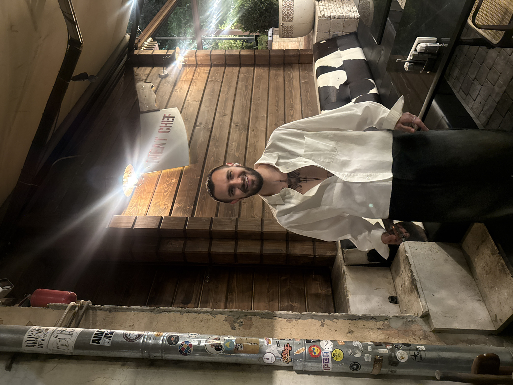 The image shows a man standing outside a building with a "MEAT CHEF" sign, possibly a restaurant in Yerevan, Armenia. The man is wearing a white shirt and dark pants. He is smiling and looking at the camera. The building has a wooden facade and a pipe with stickers running along the bottom. There is outdoor seating with a cowhide-patterned bench.