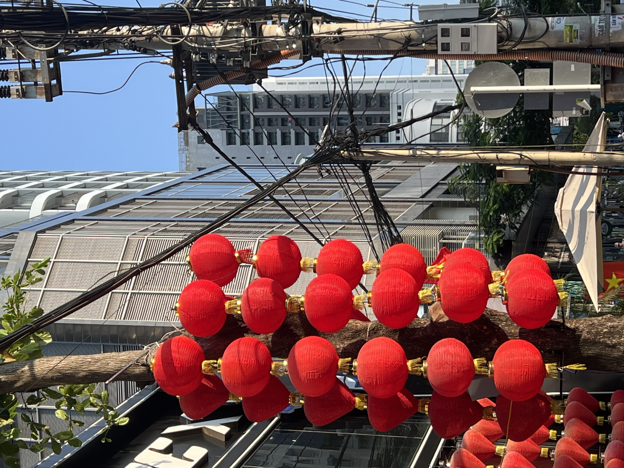 This medium, eye-level shot captures a street scene in Nha Trang, Vietnam. Red decorative lanterns are strung along a wooden branch in the foreground, reflecting on a glass surface beneath. In the middle ground, modern buildings with geometric glass facades and a taller white building are visible, partially obscured by the lanterns and overhead power lines. Power lines are highly prevalent in the upper portion of the image, crisscrossing against a bright blue sky. Also, partially visible, on the right side of the image is a Vietnamese flag.