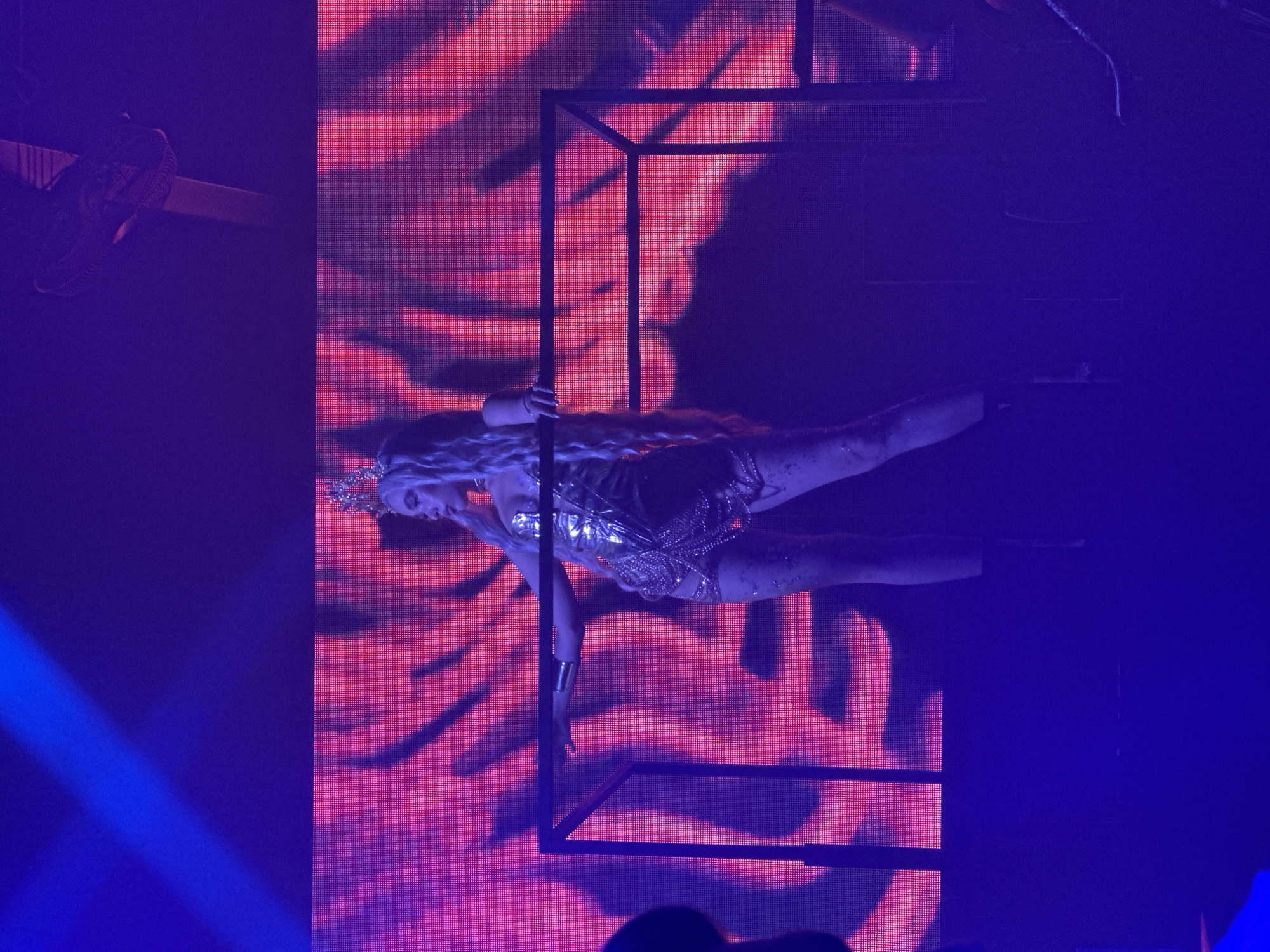 A person wearing a golden crown and a shimmering gold outfit stands on a raised platform, leaning on a railing. The background is a large screen displaying a vibrant pink and red abstract pattern. The lighting is predominantly purple and blue, with a bright blue light beam visible in the upper left corner. The scene appears to be a live performance or event, possibly a concert or a nightclub. The location is the city of Sochi, Russia.
