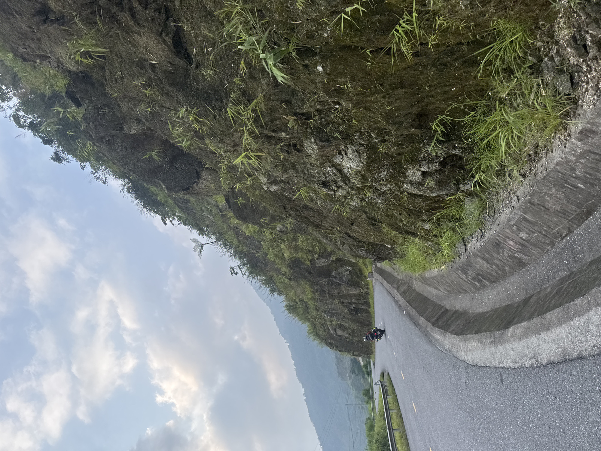 The image captures a scenic view of a paved road winding through a mountainous landscape in Tân Quang Commune, Vietnam. A dark motorcycle, with a rider, is visible on the asphalt road in the mid-ground, traveling away from the viewer. The road appears dry and has faint yellow markings on the left side, with a metal guardrail partially visible further along.
To the right of the road, a multi-tiered concrete drainage ditch or barrier separates the road from a very steep, rugged mountainside. This mountainside is primarily dark, rocky earth and stone, heavily overgrown with a variety of green vegetation, including sparse clumps of grass, ferns, and small bushes clinging to its surface. The texture of the rock and the individual plants are discernible.
In the background, beyond the immediate mountainside, multiple layers of hazy, blue-grey mountains stretch into the distance, indicating a vast and remote mountainous region. The sky occupies the upper left portion of the image, showing a mix of light blue with scattered white and grey clouds. The overall lighting suggests daytime, possibly early morning or late afternoon, with diffused light filtering through the clouds. The scene conveys a sense of peaceful travel through a natural, untamed environment. There is no visible text.