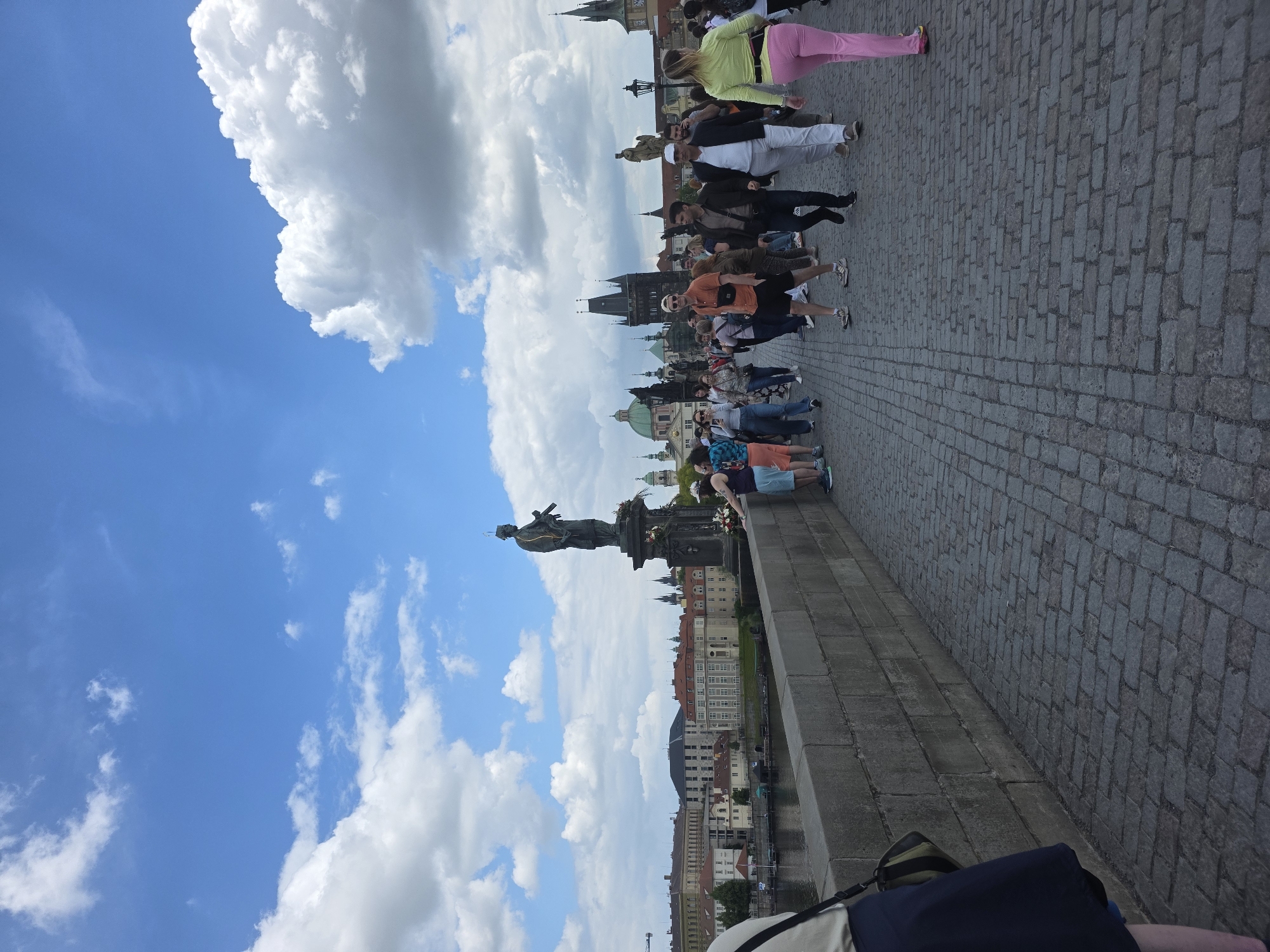 The image depicts a crowded pedestrian walkway, likely a bridge, in what appears to be a European city. The walkway is paved with cobblestones. People are walking along it. A stone wall runs along the right side of the image, and beyond it, buildings and the waterfront are visible. A statue stands on the left side, silhouetted against a cloudy sky. The image is taken at an angle, with the sky and clouds taking up a significant portion of the left side. The buildings appear to be historical, with varied architectural styles.