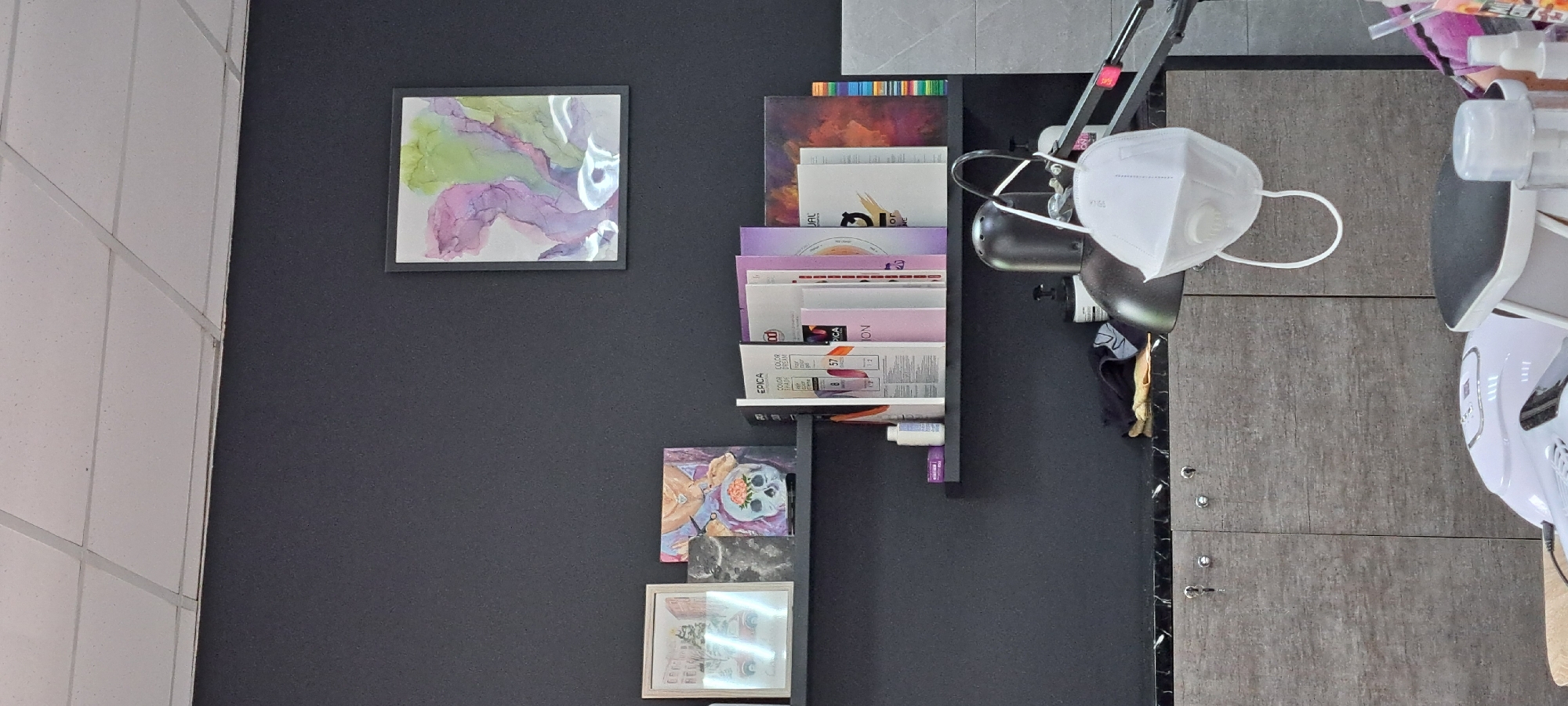 The image shows a wall with a dark gray textured finish in what appears to be a salon or beauty establishment in Yessentuki, Russia. On the wall, there are two shelves. The upper shelf holds three framed artworks. The leftmost artwork is a framed print depicting a winter scene with a red truck and trees. Next to it is a small, dark framed piece, and to its right is a larger framed artwork of a skull with flowers on top of it. The right side of the upper shelf displays a framed abstract painting with swirling purple, green, and pink colors.
The lower shelf holds several items, including bottles and a stack of what appear to be swatch books or educational materials for hair coloring, with brands like "EPICA" and "COLOR" visible. A small purple box and a white bottle with a blue label are also present on this shelf.
Below the shelves, a dark marble-like countertop runs horizontally. On this countertop, various objects are visible: a black lamp, a white face mask hanging from a stand, a black cloth, and a bottle with a black pump dispenser. Further down, there are cabinets with a gray wood-grain finish, each featuring a small silver handle and what appears to be a key lock. In the foreground, a white UV nail lamp and other salon equipment are partially visible, suggesting the setting is a functional workspace. The ceiling is composed of white acoustic tiles.