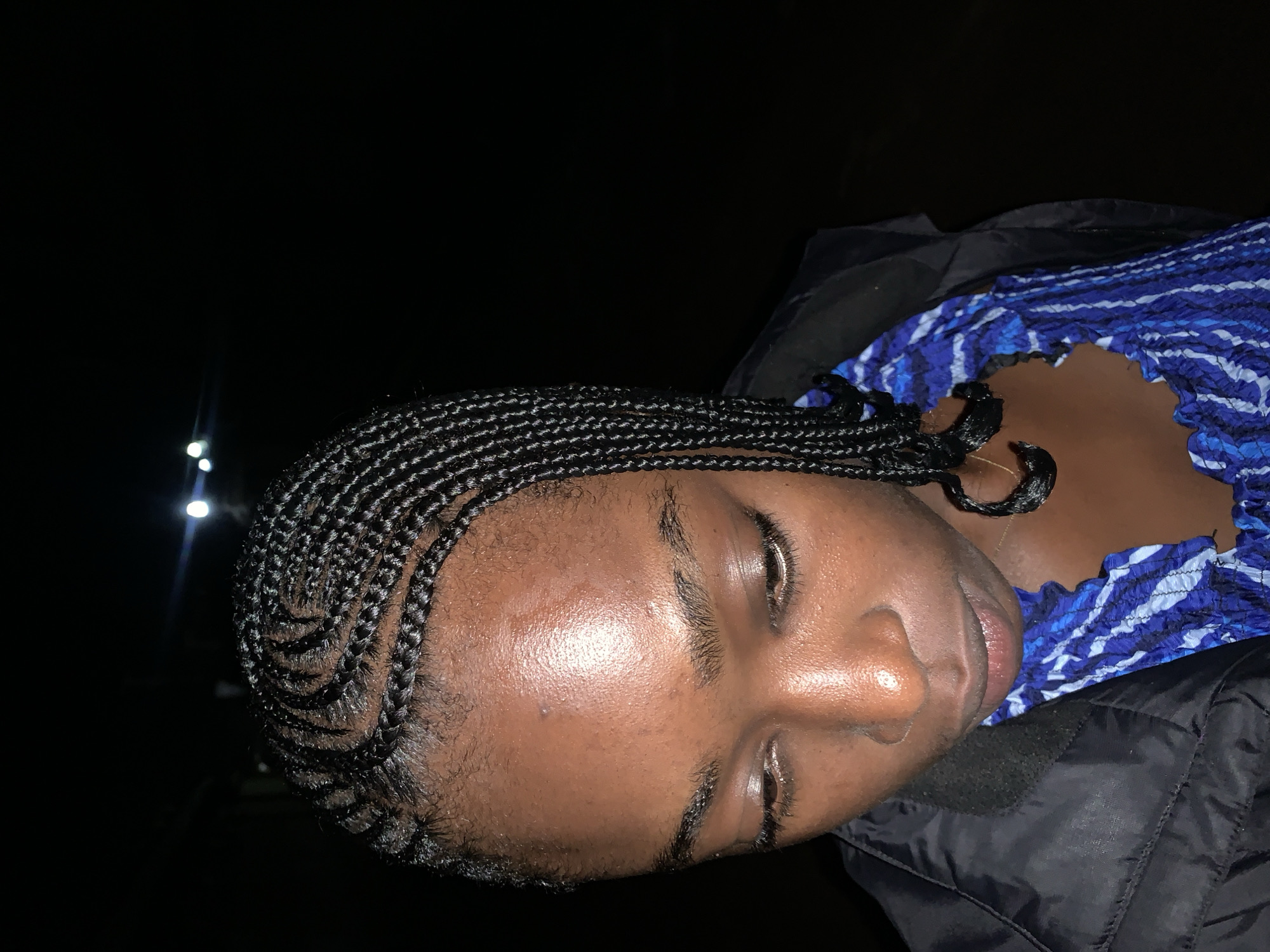 The image shows a close-up of a young Black woman from the chest up, wearing a black puffer jacket over a blue and white patterned top. Her hair is styled in small, tight braids that are neatly arranged and parted from her forehead to the back of her head. Her gaze is directed downwards, with her eyes slightly closed. The lighting suggests it is nighttime, with a dark background and a few distant lights visible. The specific location in Rafin Par, Nigeria, is not discernible from the image itself.