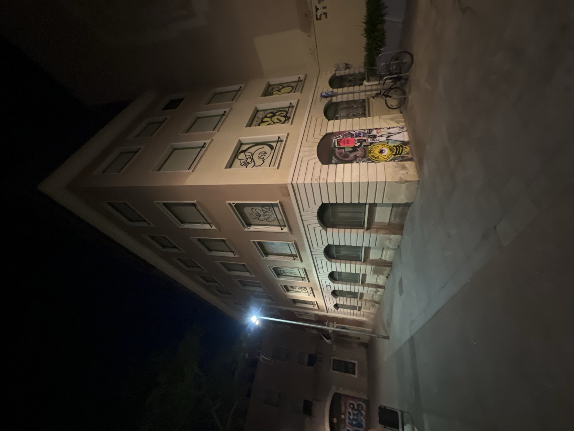 This image depicts a street corner at night in Barcelona, Spain. A four-story building with a light-colored facade dominates the scene, its windows on the upper floors are uniformly rectangular and appear to be unlit. The ground floor features arched doorways and windows, some of which are covered with graffiti. There is a streetlamp illuminating the street in front of the building, casting shadows. To the left of the building, another building with a darker facade and graffiti is visible. In front of the building on the right, there is a bicycle rack with two bicycles. The ground is paved with concrete or stone. The overall atmosphere is urban and somewhat gritty, with evidence of street art and the quiet of the night.