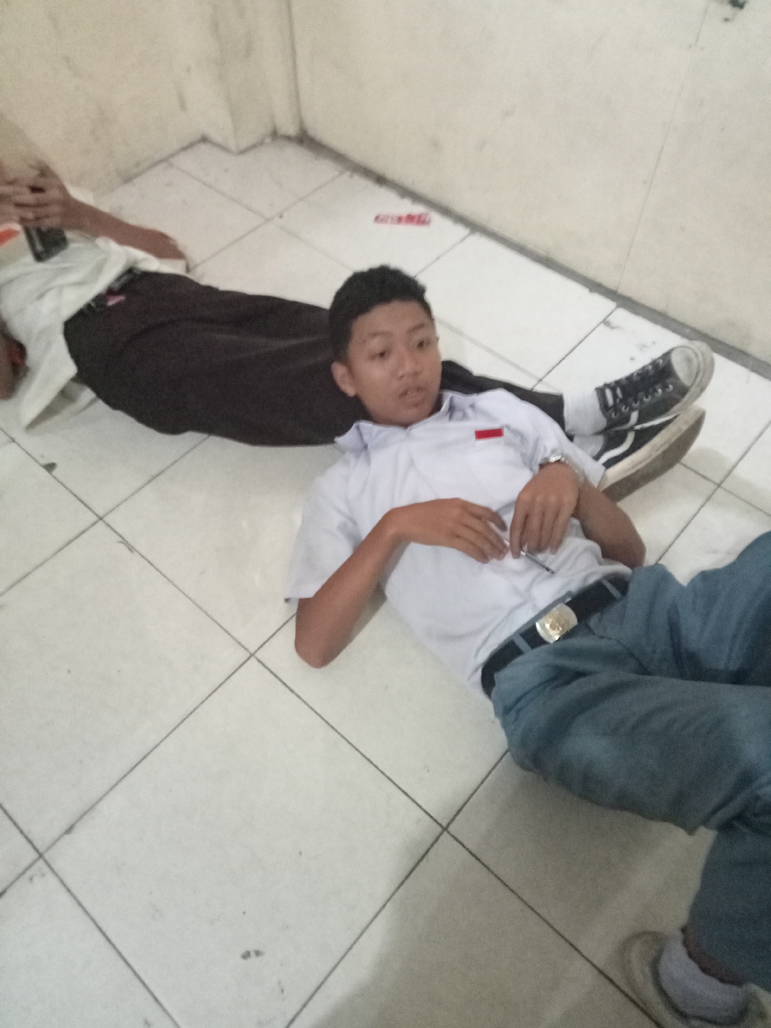 Two young males in school uniforms are lying on a tiled floor, likely indoors. The male in the foreground is wearing a white short-sleeved shirt with a red rectangular patch on the left breast, a black belt with a gold buckle, and blue trousers. He is lying on his back, with his right arm resting on his abdomen and holding a pen. His left arm is bent at the elbow. He is wearing black sneakers with white laces and soles. The second male, in the background, is wearing a white short-sleeved shirt and dark trousers. He is also lying on the floor, partially visible. The environment appears to be a school hallway or room, with a light-colored wall and a cream-colored tiled floor with dark grout lines. The lighting is dim. There are no distinct landmarks in the image that would confirm the location in Secang, Indonesia.