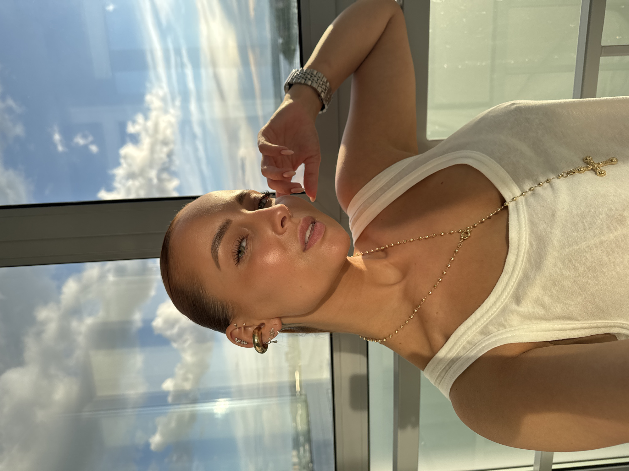 A woman with light skin is shown in a close-up, slightly tilted shot in front of a window. She is wearing a white tank top and jewelry, including a watch, a gold necklace with a cross pendant, and multiple earrings. She has her hand near her face. Outside the window, there is a view of a sky with clouds and what appears to be a cityscape. Based on the location information provided, this cityscape is of Izhevsk, Russia. There is no interaction apparent in this image.
