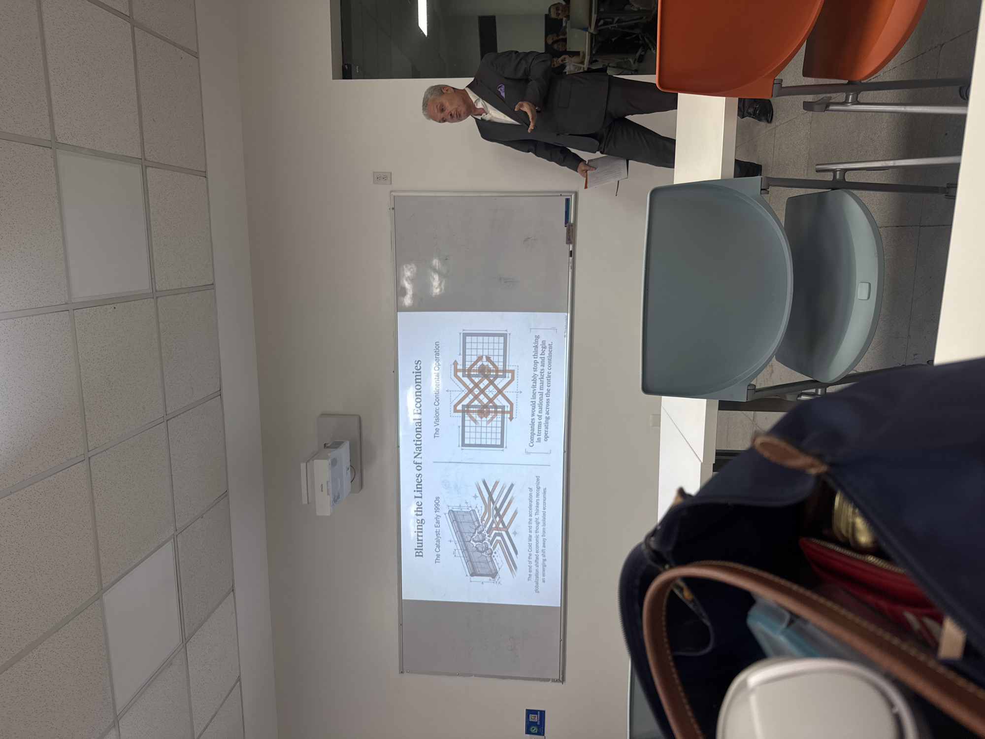 The image depicts an indoor presentation or lecture taking place in a modern classroom or conference room in Puebla, Mexico. A man, likely the presenter, stands to the right of a whiteboard, gesturing with his left hand while holding papers in his right. He is dressed in a dark suit, white shirt, and a purple pocket square, appearing poised and engaged.
A projector, mounted on the ceiling to the left, casts a presentation slide onto the whiteboard. The slide is titled "Blurring the Lines of National Economies." Below the title, two sections are displayed: "The Catalyst: Early 1990s" and "The Vision: Continental Operation." The text explains: "The end of the Cold War and the acceleration of globalization shifted economic thought. Thinkers recognized an emerging shift away from isolated economies." and "Companies would inevitably stop thinking in terms of national markets and begin operating across the entire continent." The slide also includes illustrative diagrams of interconnected lines and grids.
The room features a white dropped ceiling with acoustic tiles. Several modern chairs, including two orange chairs and two grey chairs, are arranged around a white table in the foreground and midground, suggesting an audience is present or has been. In the immediate foreground, a dark blue bag is partially visible, containing personal items such as a red wallet, a gold object, and a light blue container. The setting appears well-lit by artificial lighting, typical of a professional or educational environment.