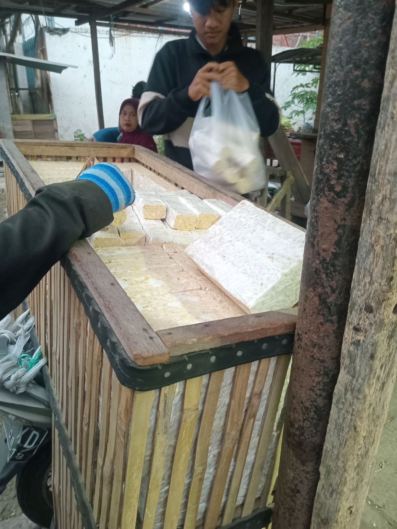 The image depicts an outdoor scene, likely in Kwangsan, Indonesia, where a person wearing a black jacket and blue and white striped gloves is reaching into a large, wooden, slatted crate. Inside the crate are numerous rectangular blocks of tempeh, some of which are partially covered by a plastic bag held by another person. A woman wearing a maroon hijab is visible in the background, looking towards the crate. To the right of the crate, two large, rough-textured logs stand vertically. A motorcycle license plate with the characters "XD" and the number "19" is partially visible in the lower left corner. The overall environment appears to be rustic, possibly a market stall or a food preparation area, with a covered structure overhead and glimpses of vegetation in the background.