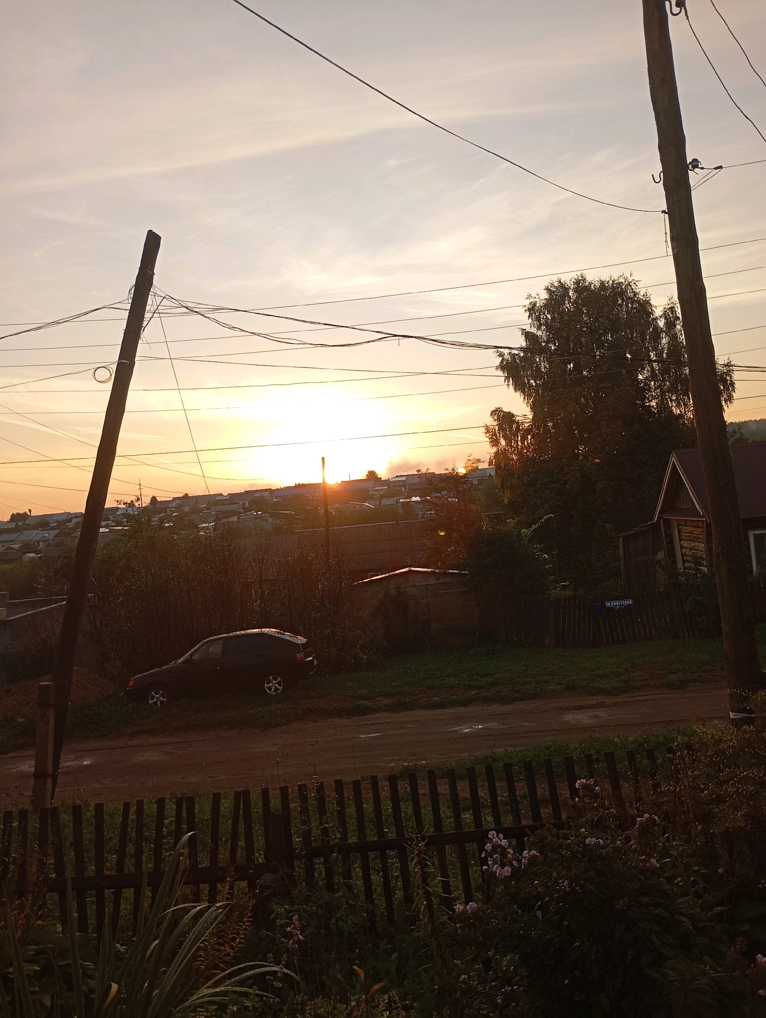 The image depicts a rural landscape at sunset, likely in the city of Foky, Russia. In the foreground, a wooden fence made of vertical planks separates a grassy area with some flowering plants from a dirt road. A dark-colored hatchback car is parked on the grassy area to the left of the road. Behind the car and the road, there is a densely vegetated area with trees and bushes. In the middle ground, a collection of houses, characteristic of a village or small town, is spread across a gentle slope. The houses have dark roofs and are of varying sizes. In the background, a large deciduous tree stands prominently, silhouetted against the sky. The sky is filled with the warm hues of a sunset, with the sun low on the horizon, casting a golden glow and some light clouds. Utility poles with numerous electrical wires are visible, stretching across the upper portion of the image, indicating the presence of infrastructure in the area. The overall impression is one of a tranquil, early evening scene in a rural Russian settlement.