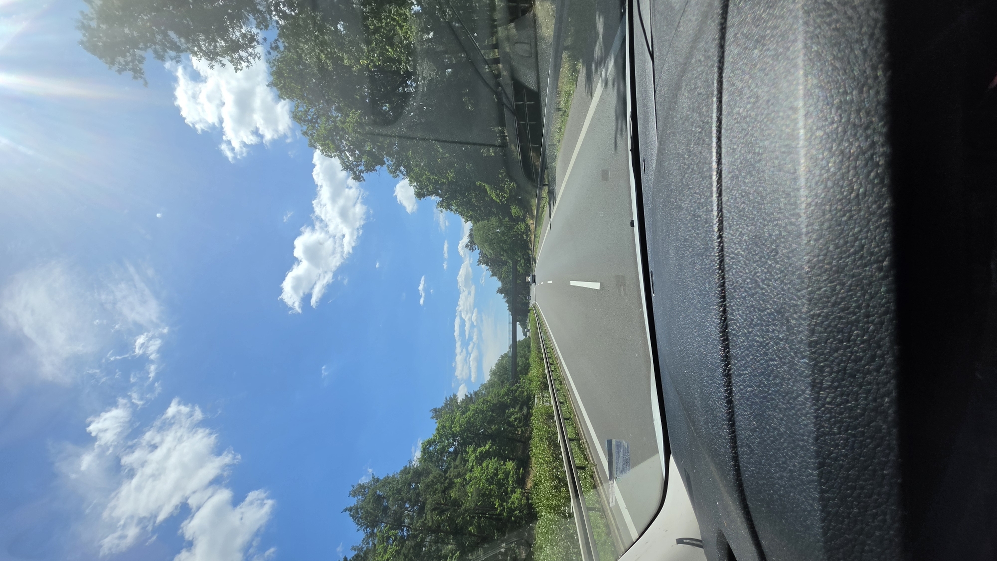 The image depicts a view from inside a vehicle, looking out through the windshield onto a two-lane road. The road appears to be a highway, bordered by a guardrail and lush green trees on both sides. The sun is shining brightly, casting rays of light that create a lens flare in the upper left corner of the image. The sky is a vibrant blue, dotted with fluffy white clouds, suggesting fair weather. The time of day is likely daytime, given the strong sunlight. The interior of the vehicle is visible on the right side of the frame, showing a textured dark gray surface, possibly part of the dashboard or door panel. A thin white line is visible on the road, indicating a lane marker. In the distance, the road curves slightly and there appears to be a bridge or overpass. The overall scene evokes a feeling of travel and a pleasant, sunny day. There is no visible text in the image. The location context provided is Calau, Germany.